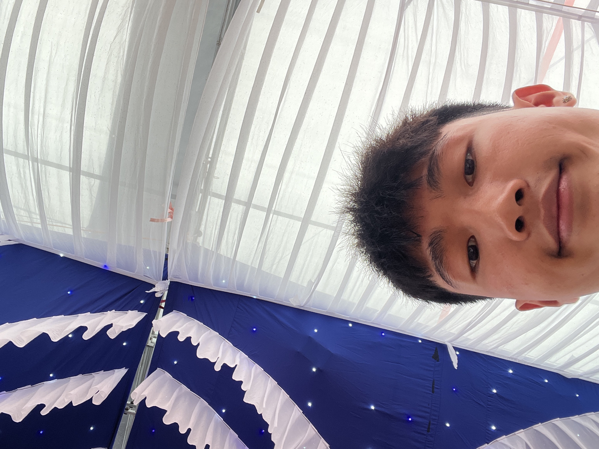 The image is a selfie taken from a low-angle perspective, looking upwards at the decorated ceiling of a tent or canopy. A young man, likely of East Asian descent, is featured prominently in the top right portion of the frame, appearing upside down from the viewer's orientation. He has short dark hair, an earring in his visible right ear, and looks directly into the camera with a neutral or slightly smiling expression. His eyes, nose, and part of his mouth are clearly visible.
The tent's ceiling is adorned with two primary fabric types. The upper section, where the man's head is largely positioned, consists of translucent white fabric panels with visible vertical folds, suggesting diffuse natural light or bright artificial lighting from above. The lower section is made of a deep royal blue fabric, embellished with decorative white ruffled fabric strips forming a wave-like or scalloped pattern. Numerous small, star-like lights, both white and blue, are embedded within the blue fabric, creating a festive atmosphere. Structural poles or framing are visible where the different fabric sections converge.
The scene depicts the interior of a decorated event tent, indicative of a celebration or gathering such as a wedding or party. The location context is Tay Son Commune, Vietnam. There is no visible text in the image.