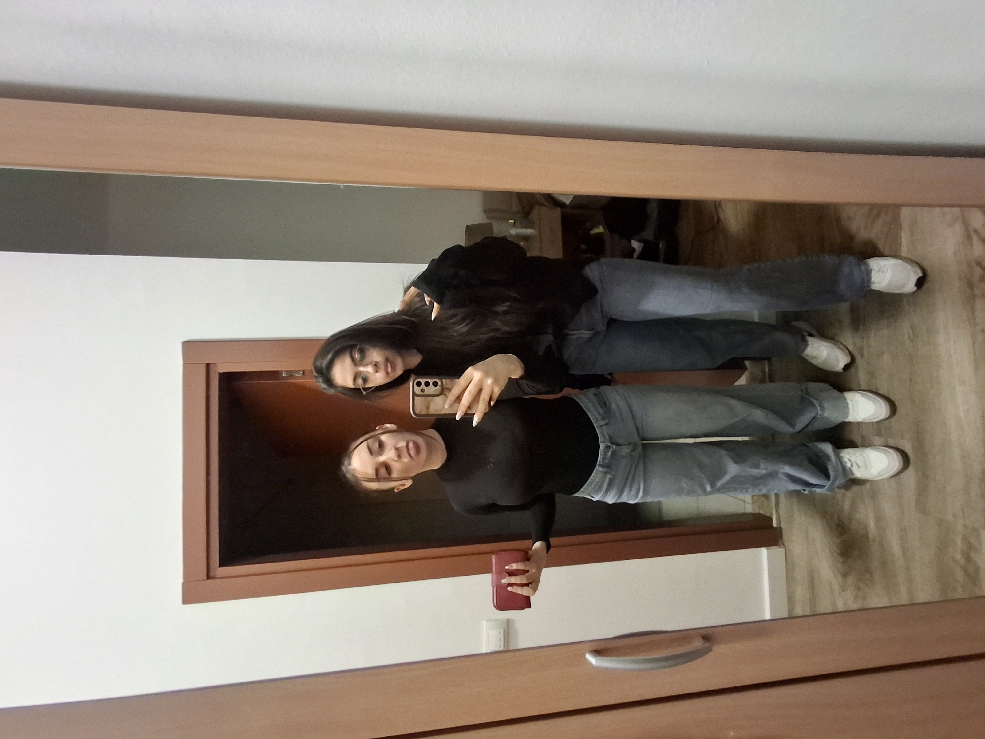 Two young adult females are reflected in a full-length mirror within a residential interior, specifically in the Vozdovac Urban Municipality, Serbia. The scene depicts a selfie being captured.
The female on the left in the reflection has light skin, brown hair pulled back, and is wearing a black long-sleeved turtleneck top and light-wash, wide-leg blue jeans cuffed at the ankles, paired with white sneakers. She holds a red rectangular object, possibly a wallet or clutch, in her left hand and extends her tongue in a playful expression. Her right hand, partially visible, has long, light-colored fingernails.
The female on the right in the reflection has darker skin, long dark hair, and wears clear-framed glasses. She is dressed in a dark, long-sleeved top and dark-wash blue jeans, with white sneakers featuring dark soles. She is holding a smartphone with a patterned case in her right hand, capturing the mirror reflection. Her left hand is raised near her chest, also displaying long, light-colored fingernails. Her facial expression is neutral as she looks towards the phone.
The immediate environment visible in the reflection includes light-colored walls, a light brown wooden door frame and door, and light brown wooden laminate flooring. To the far left, a light brown wooden wardrobe or cabinet with a silver handle is partially visible. In the background reflection beyond the individuals, a wooden piece of furniture, possibly a bedside table with a lamp, can be faintly discerned.