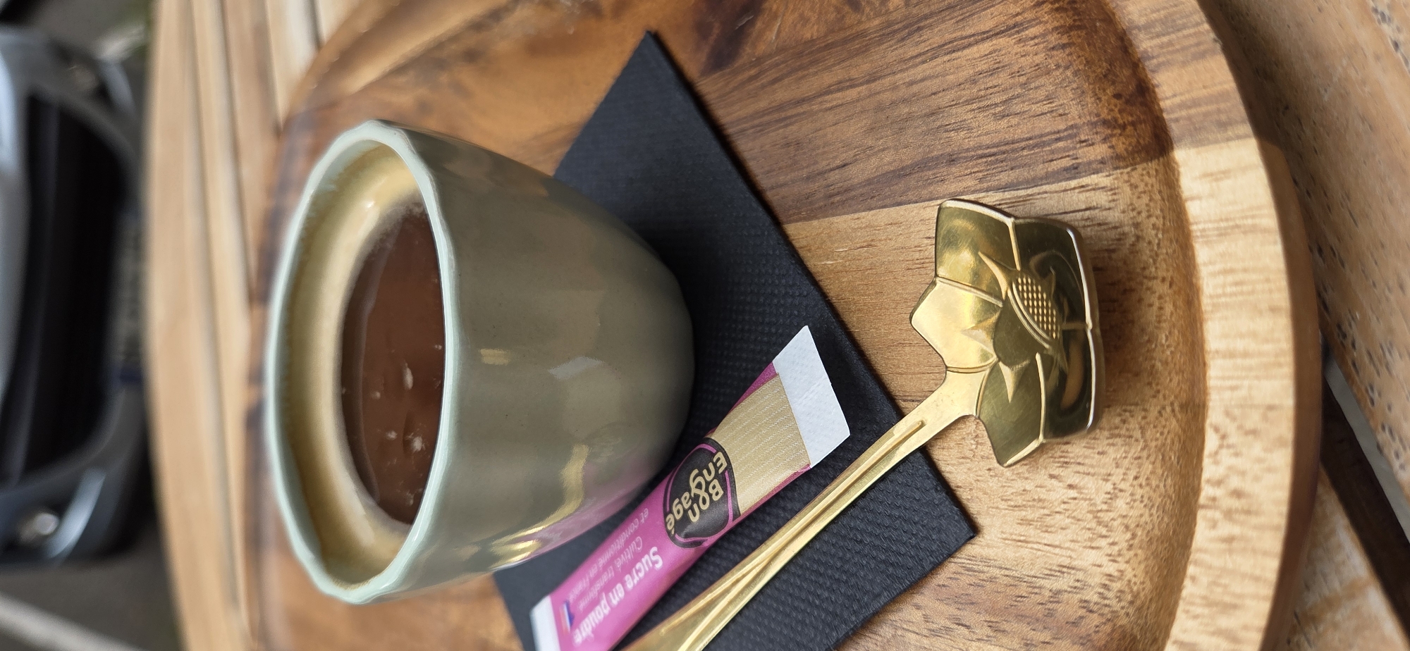 This close-up, vertical, eye-level shot captures a small cup of dark coffee or espresso, filled to the brim, resting on a black square napkin. Beside the cup, a pink and gold packet of sugar, labeled "Sucre en poudre" and "Engage," lies diagonally. A gold spoon with an ornate, flower-like handle is positioned in front of the sugar packet and napkin, its bowl angled slightly towards the viewer. The setting appears to be an outdoor cafe or restaurant, with a warm-toned wooden table surface visible beneath the napkin and a blurred background that suggests a street or urban environment, possibly with a hint of a car. The image is likely taken at an outdoor table, possibly in a pedestrian zone or near a street. The details are consistent with an outdoor dining experience in a city like Strasbourg, France, known for its charming cafes and bistros.