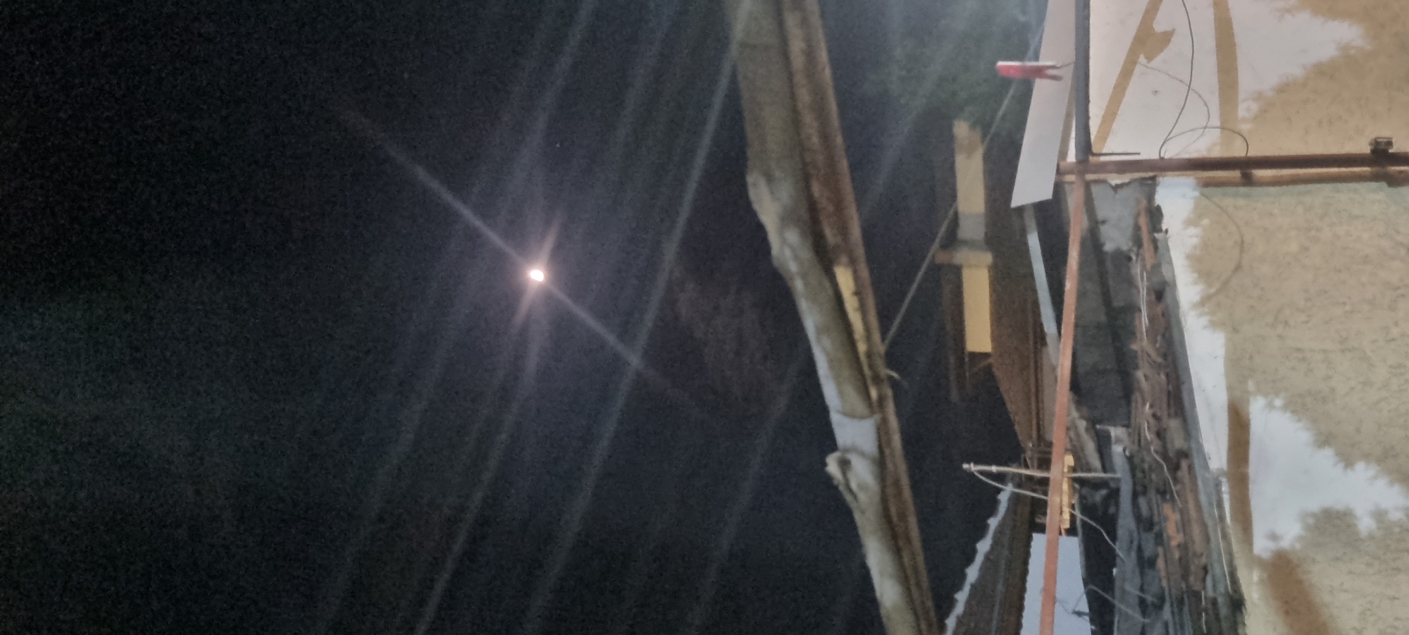 The image shows a night scene. A bright light, possibly the moon or a distant street light, is visible in the dark sky on the left side. To the right, a cluttered scene of building materials, what appears to be part of a building or structure under construction or repair, with a mix of wood, metal, and possibly some electrical wiring. The scene is grainy and lacks fine detail due to the low light conditions. There are no visible people. The location appears to be an urban or semi-urban area in an area of Unknown, Unknown.