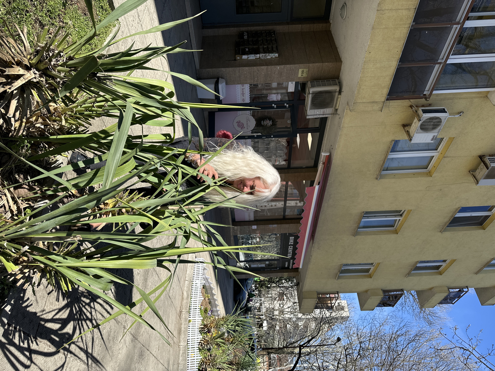 A woman with long, white hair is partially obscured by the foliage of a yucca plant on a sidewalk in Sofia, Bulgaria. She appears to be looking down. In the background is a building with a yellow facade and several windows. A commercial establishment, possibly a "Bike Center," is visible behind her. A white picket fence runs along the sidewalk in the mid-ground. The sky is blue, and trees are partially visible in the background. The image is taken during the day with bright sunlight.