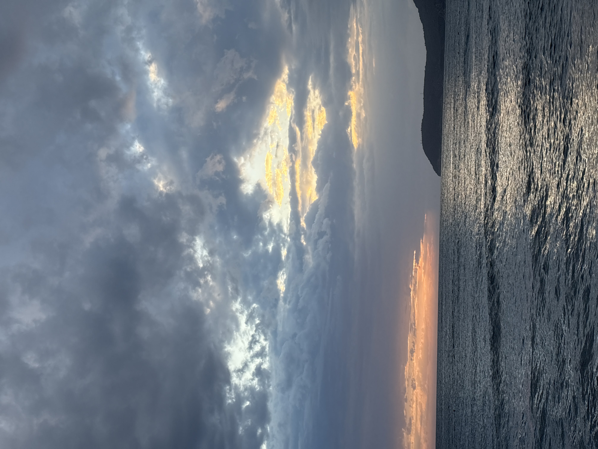 The image captures a seascape at Bar, Montenegro. The scene is divided vertically, with the sky dominating the left side and the sea on the right. In the sky, there are dark, turbulent clouds illuminated with yellow and orange light near the horizon. The sun appears to be setting or rising. Adjacent to the sea, a dark mass of land, likely a mountain, is visible. The sea is rippled, with reflected sunlight. No people are visible.