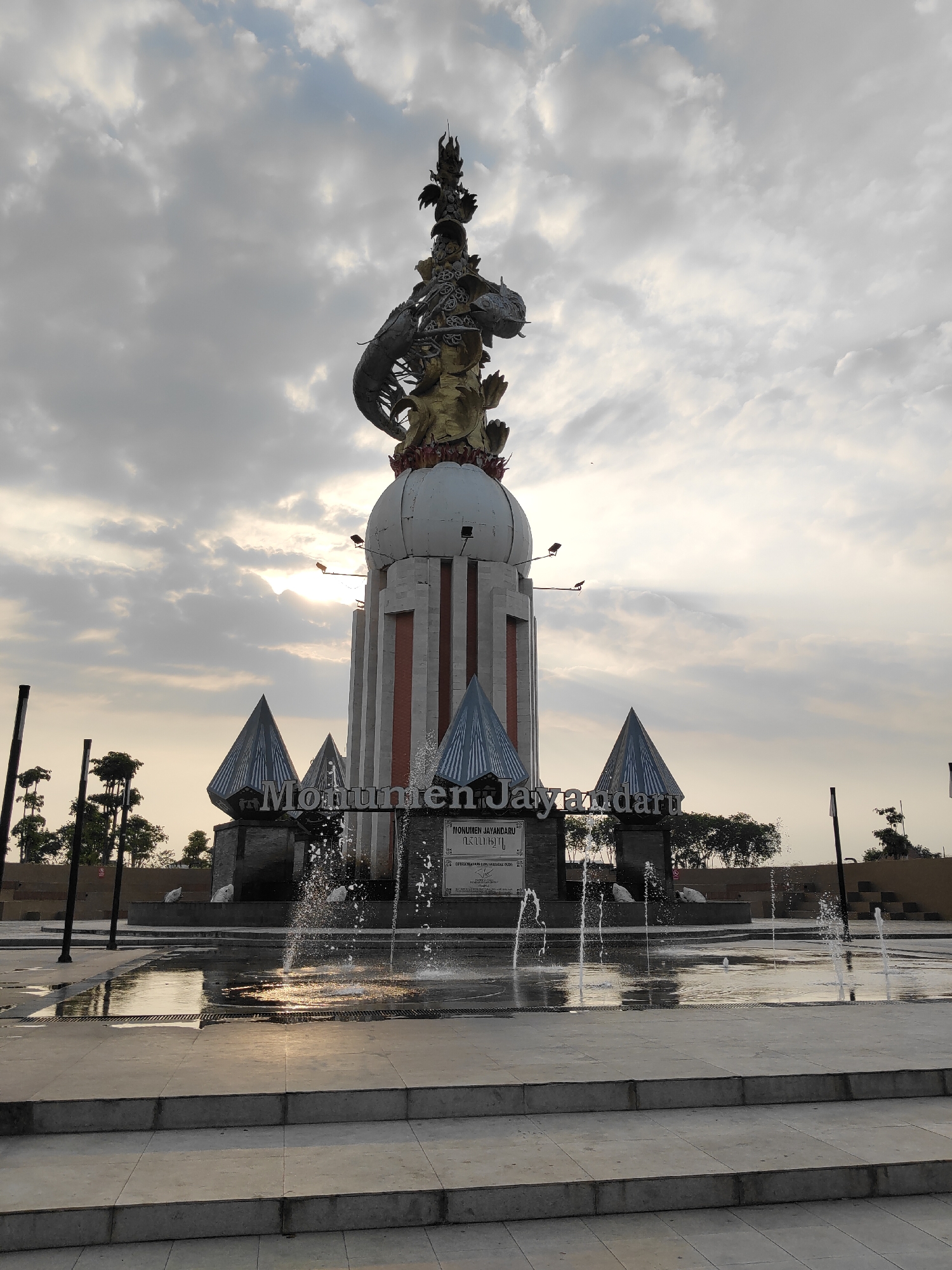 This image depicts the Jayandaru Monument in Sidoarjo, Indonesia, under a cloudy sky with the sun partially visible behind the clouds. The monument is a tall, ornate structure with a white domed base and a complex, multi-tiered top featuring golden and metallic figures, including what appears to be a dragon or serpent. The base of the monument is surrounded by three pyramid-shaped roofs and a fountain with water spraying into a shallow pool. A sign in front of the monument reads "MONUMEN JAYANDARU". The foreground shows a wet paved area with steps leading down to the fountain. There are no people visible in the image. The environment suggests an outdoor public space.