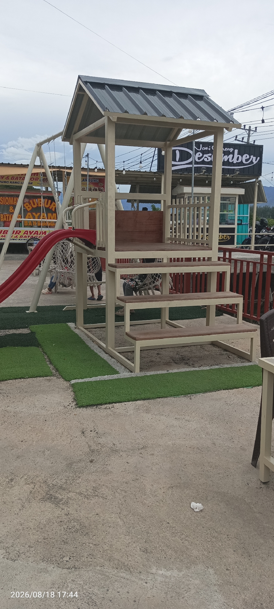 The image depicts an outdoor scene, likely a recreational area adjacent to a food establishment, in Cotcut, Indonesia. The focal point is a beige metal playground structure with a gray metal roof. This structure features two wooden benches integrated as steps, leading up to a wooden platform. To the left of the structure, a red slide curves downwards, and a white net swing is suspended.
In the background, several food stalls or small shops are visible, with signage indicating food items. One prominent sign reads "Nasi Goreng Desember" (Fried Rice December). String lights are hung above the stalls. Beyond the stalls, a mountainous landscape is visible under a cloudy sky.
Several individuals are present in the scene, though their interactions are not clearly discernible. Two people, one wearing a floral shirt and the other a dark shirt, are seated on chairs or benches within or near the playground structure. Another individual, wearing a light-colored top and patterned skirt, is seen sitting on a swing. A person in a dark shirt and shorts is standing near the playground.
The ground is a mixture of concrete and artificial turf, with rectangular sections of green turf laid out in a patterned fashion. A crumpled white tissue is visible on the concrete floor in the foreground. The overall atmosphere suggests a casual, possibly commercial, outdoor space. The timestamp "2026/08/18 17:44" is overlaid on the bottom left of the image.