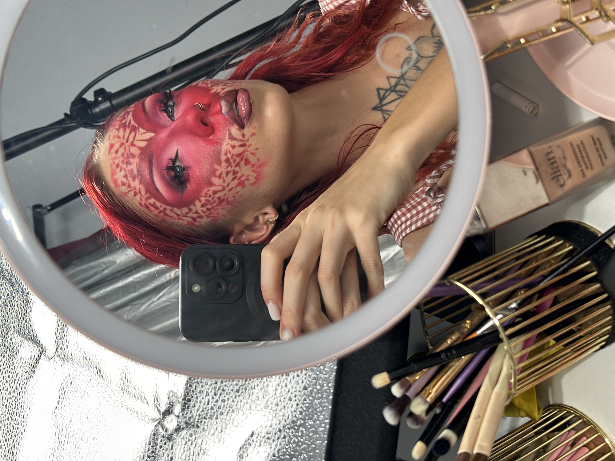 A woman with vibrant red hair and elaborate red and white face makeup, resembling a floral pattern, is reflected in a ring light mirror. She is taking a selfie with her phone, which is also visible in the reflection. The makeup includes dramatic eye makeup, adorned eyebrows, and piercings on her nose and lip. She wears a gingham shirt. In the background, reflected in the mirror, are makeup brushes in a gold holder and a bottle of "elian." foundation. The lighting suggests an indoor setting, likely a makeup studio or dressing area, possibly during the day. The overall mood is artistic and creative, focusing on the transformation and application of makeup.