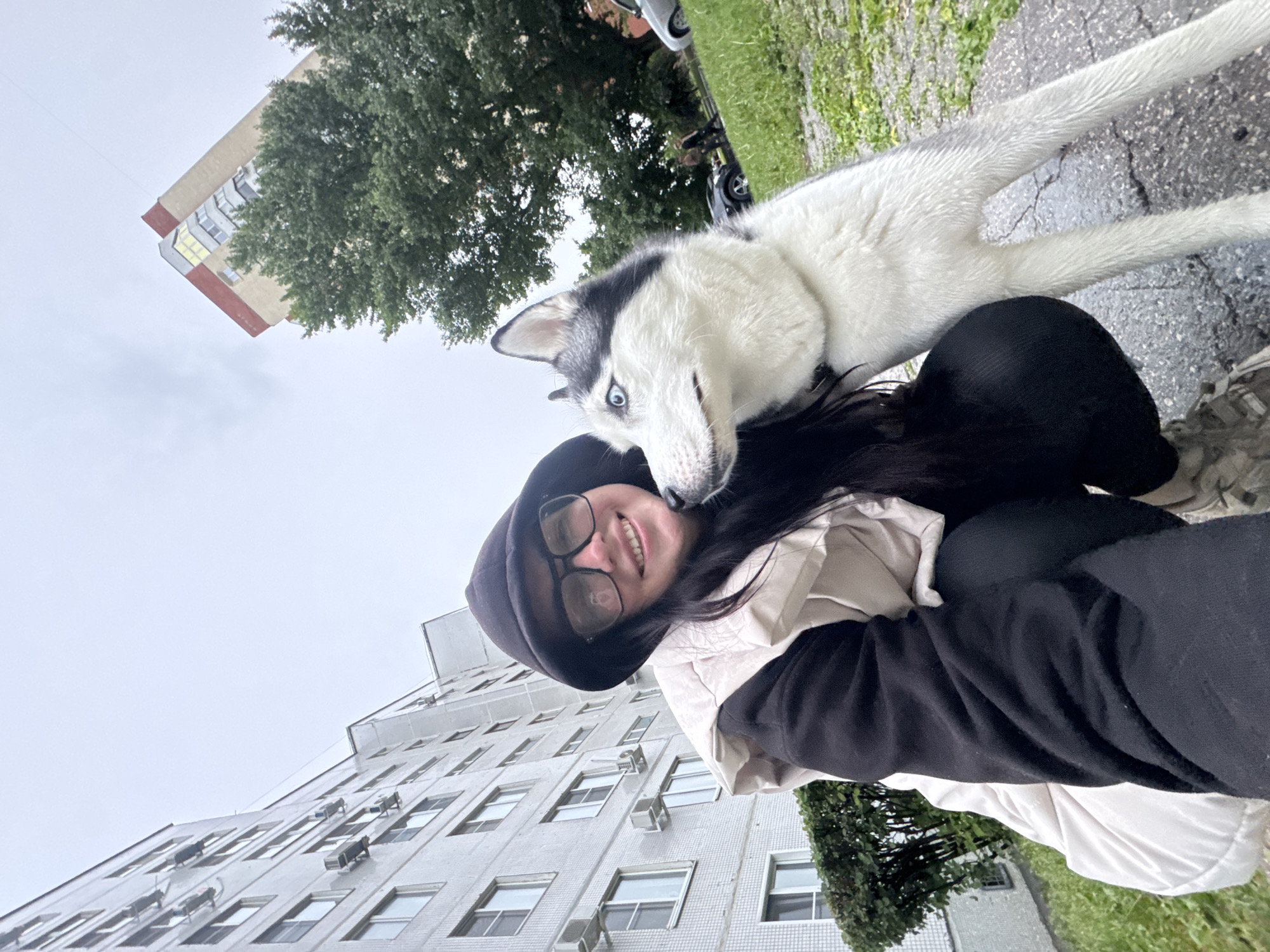 The image captures a cheerful outdoor interaction between a young woman and a black and white husky dog. The woman, with dark hair, glasses, and wearing a black hoodie over a light-colored jacket, smiles broadly, looking towards the camera. The husky, with striking blue eyes and a distinctive black and white coat, has its head leaning against the woman's cheek, appearing affectionate or curious.
They are situated outdoors, with an overcast sky dominating the upper part of the background. A large, multi-story building, possibly an apartment complex, with numerous windows and visible air conditioning units, stands behind them. The building is light-colored, likely white or cream. Green foliage from trees and patches of grass are visible on the ground, alongside a cracked, dark paved surface. The scene suggests a daytime setting, possibly under cloudy weather conditions. The woman's joyful expression and the dog's gentle posture convey a warm and happy moment. No discernible text is visible in the image.