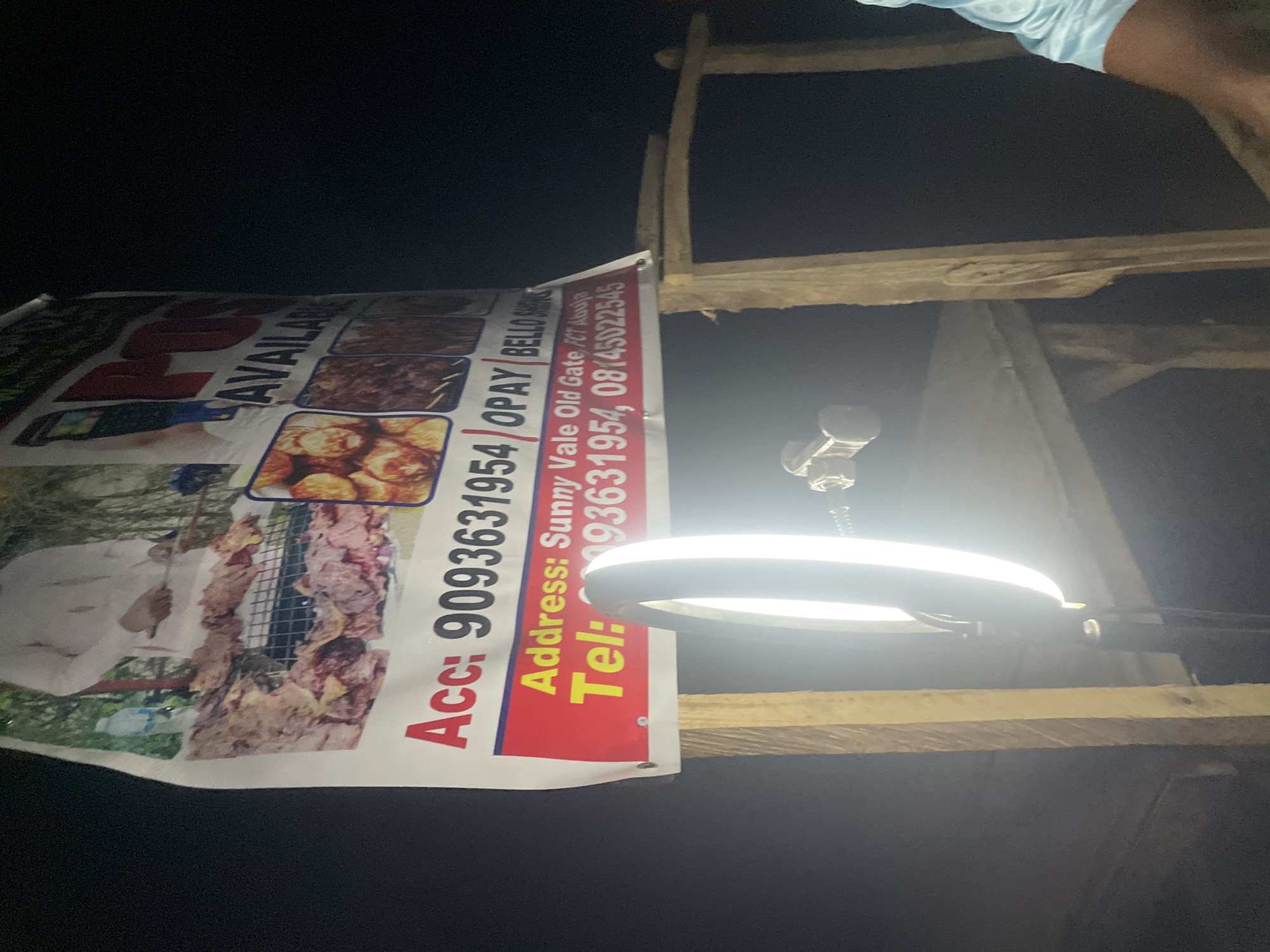 The image captures a nighttime scene, likely outdoors or in a dimly lit indoor setting, illuminated primarily by an artificial ring light positioned on the right. A large, vertically oriented banner or poster is prominently displayed on the left, advertising food items, specifically what appears to be skewered meat or kebabs, alongside other roasted or fried dishes.
A person's arm and shoulder, wearing a light blue garment, are partially visible at the top right corner. On the banner itself, a person in a white garment is depicted alongside the food. Above the ring light, a small camera or smartphone is mounted, suggesting an activity of recording or photography, possibly for advertising the food products.
The banner contains visible text providing business information:
- "AVAILAN" (likely part of "AVAILABLE")
- "Acc: 9093631954/OPAY / BELLO" (suggesting an account number for the OPay payment platform and a name)
- "Address: Sunny Vale Old Gate, FC Abuja" (indicating a location within Abuja, Nigeria)
- "Tel: 93631954, 08145022545" (providing contact phone numbers).
Wooden planks or scaffolding form part of a structure, possibly supporting the banner or acting as a stall framework. The overall scene, set in Galadimawa, Nigeria, depicts a food vendor operating at night, utilizing a ring light for illumination, possibly for a photoshoot or video, to promote their business.