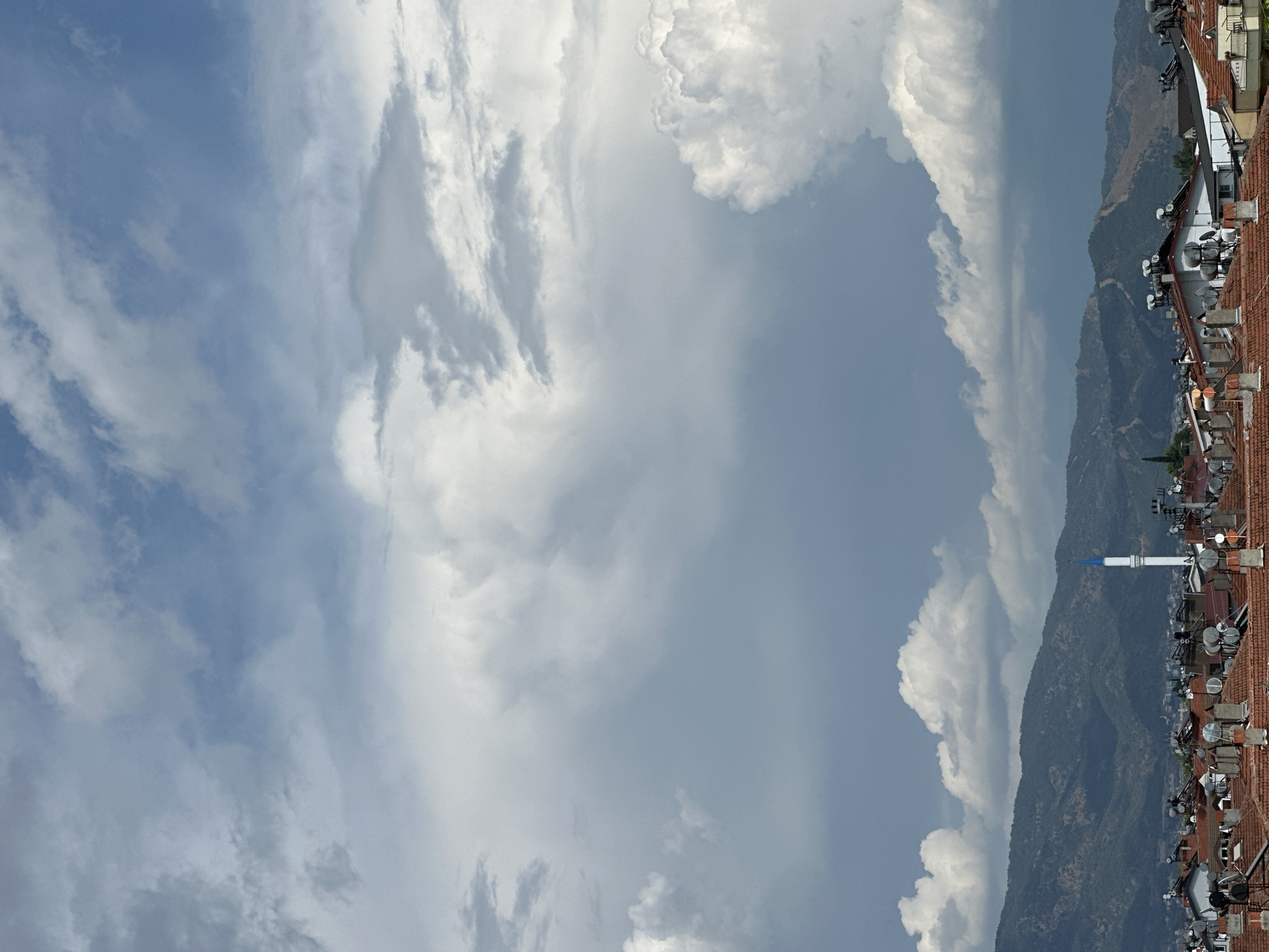 The image shows a daytime view in Fethiye, Turkey. The sky, filled with various cloud formations, takes up a significant portion of the frame. A mountain range, partially obscured by clouds, runs along the right side of the image. In the foreground, there's a view of rooftops with red tiles, numerous satellite dishes, and a white minaret with a blue top.