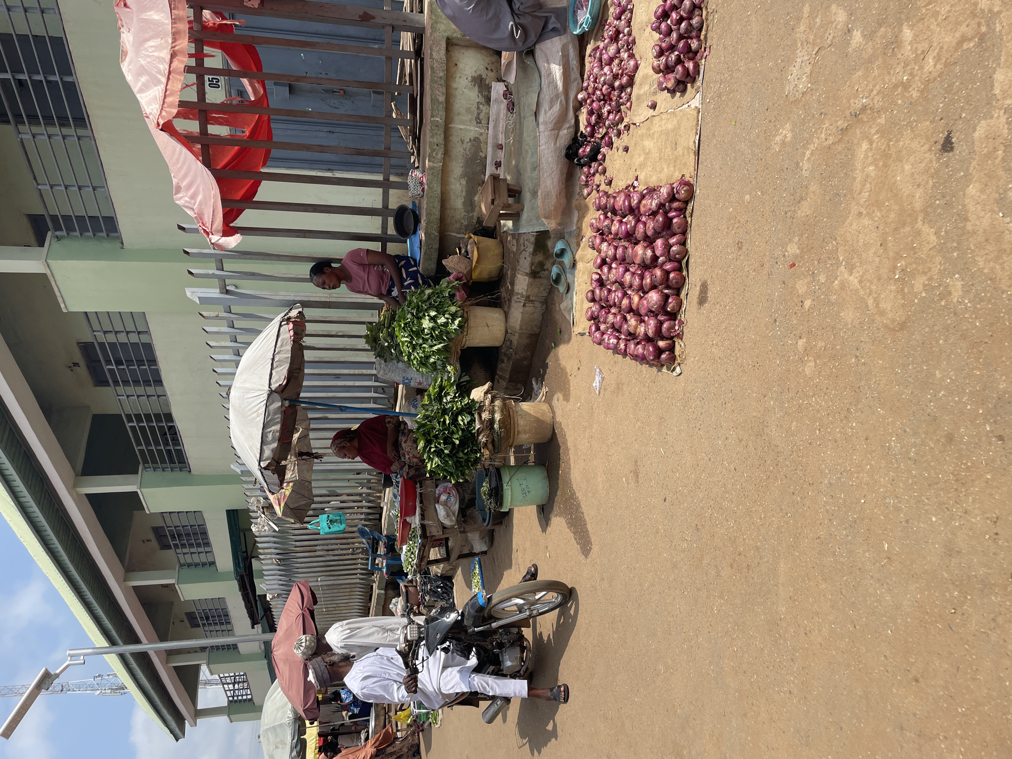 A street market scene is depicted in Akwanga, Nigeria. In the foreground, large piles of red onions are displayed on the ground, spread across two pieces of rough fabric. To the left, a man wearing a white traditional outfit and a cap rides a motorcycle. Behind him, several market stalls are set up with umbrellas providing shade. One stall features a woman in a red headscarf and traditional attire sitting next to a display of leafy green vegetables. Another stall has a woman in a pink t-shirt and a patterned skirt sitting and also with vegetables. The background shows a two-story building with a light green facade and railings on the balconies, suggesting a commercial or residential area. A metal fence separates the market stalls from the building. The ground is dusty and appears to be unpaved.