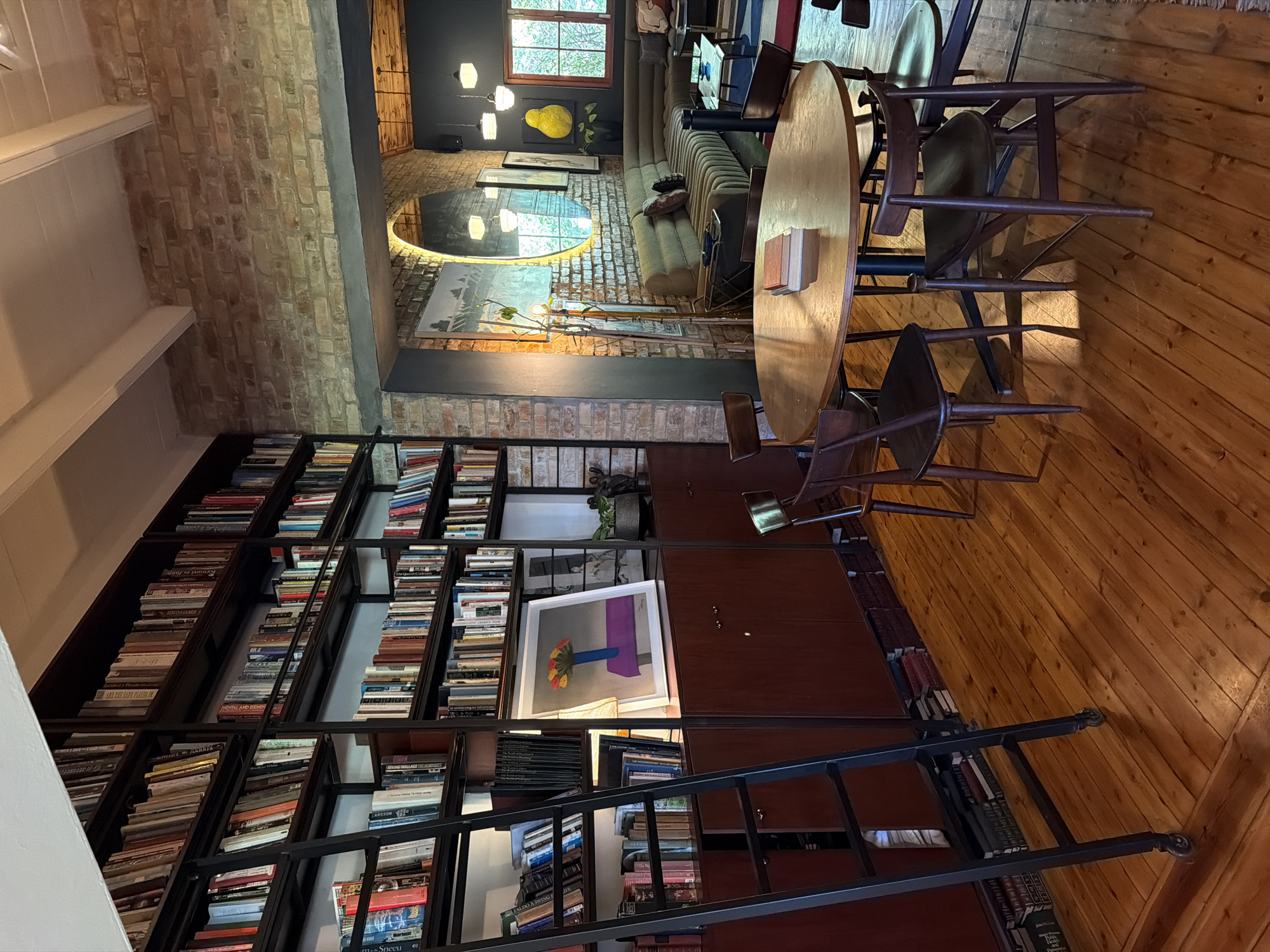 The image depicts a warm, inviting room with a prominent, floor-to-ceiling bookshelf filled with books. A black metal ladder leans against the bookshelf, suggesting accessibility to higher shelves. Below the bookshelf, dark wooden cabinets with handles are visible, some of which contain books. A framed artwork of a blue vase with flowers on a purple surface hangs on the wall.
In the foreground, a round wooden table is set with several wooden chairs. Two books are stacked on the table. The floor is made of polished wooden planks.
In the background, through an archway, a living area is visible with a large, sectional sofa and coffee tables. The walls are adorned with artwork, and a large circular mirror reflects the scene. The overall ambiance is cozy and suggests a space dedicated to reading and relaxation.
This scene is located in the city of Overstrand Nu, South Africa, likely in a residential or a hospitality setting such as a guesthouse or a library-themed cafe.