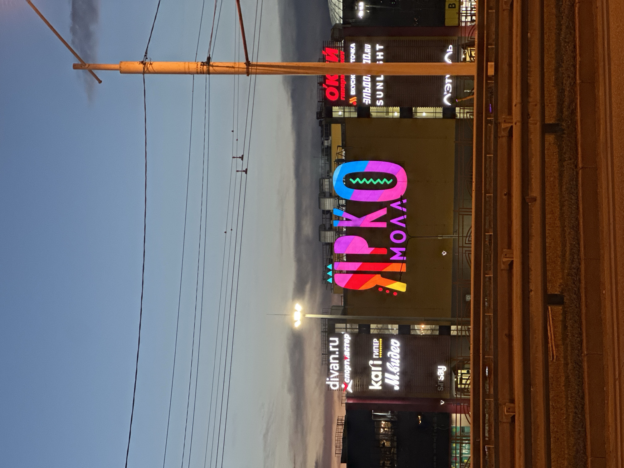 This is a vertical, eye-level photograph taken at dusk in Irkutsk, Russia, showing a street scene with a shopping mall and overhead power lines. The sky is a gradient of pale blue to soft gray, indicating the transition from day to night.
The primary subject is a large, brightly lit sign for "SIPKO MOLL" that features a colorful, abstract design resembling a guitar with a wavy pattern inside a rounded shape. Below this, the mall features several illuminated signs for various stores, including "divan.ru" (sportmaster), "kari ГИПЕР", and "M.видео". Other visible text includes "ОКИ", "ВКУСК ТОЧКА", "ЭЛЬДО 10.RU", and "SUNL BHT", "sinsay", and "Л'Эт". The mall appears to be a modern structure with large windows, some of which are illuminated from within.
In the foreground, a utilitarian yellow pole with wires stretches diagonally across the frame, suggesting the presence of streetlights or trolley lines. The wires themselves are a prominent feature, crisscrossing the sky and adding a sense of urban infrastructure to the scene. On the right side of the image, a textured, reddish-brown structure, possibly a wall or fence made of corrugated metal, occupies the right third of the frame, creating a strong vertical element.
The overall mood is somewhat subdued, characteristic of twilight. The bright lights of the signs stand out against the darkening sky, highlighting the commercial activity of the shopping center. The scene conveys a typical evening in a Russian city, with a focus on consumerism and public utilities. No people are clearly visible.