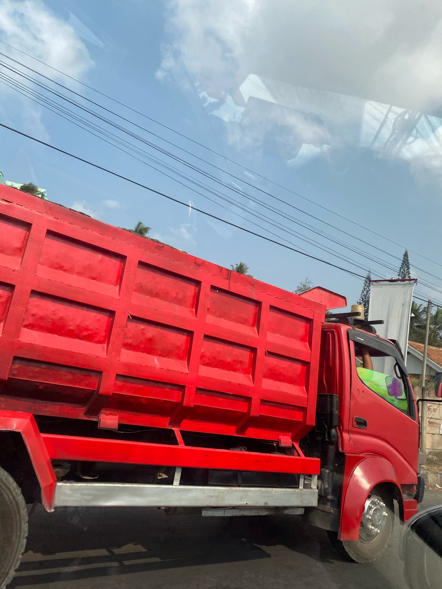 A bright red dump truck is captured in profile, moving along a paved road. The truck's bed is large and segmented, with a sturdy construction. A portion of the driver's cabin is visible, with reflections of a green and purple object on the passenger side window. The scene is set outdoors, under a bright blue sky dotted with white clouds. Several power lines stretch across the sky, and a tall evergreen tree stands in the background to the right, alongside a white banner attached to a pole. In the distance, houses with terracotta roofs can be seen. The environment suggests a location in a city or town, possibly in a tropical or subtropical region given the foliage. The specific location is identified as Bandar Lampung, Indonesia.
