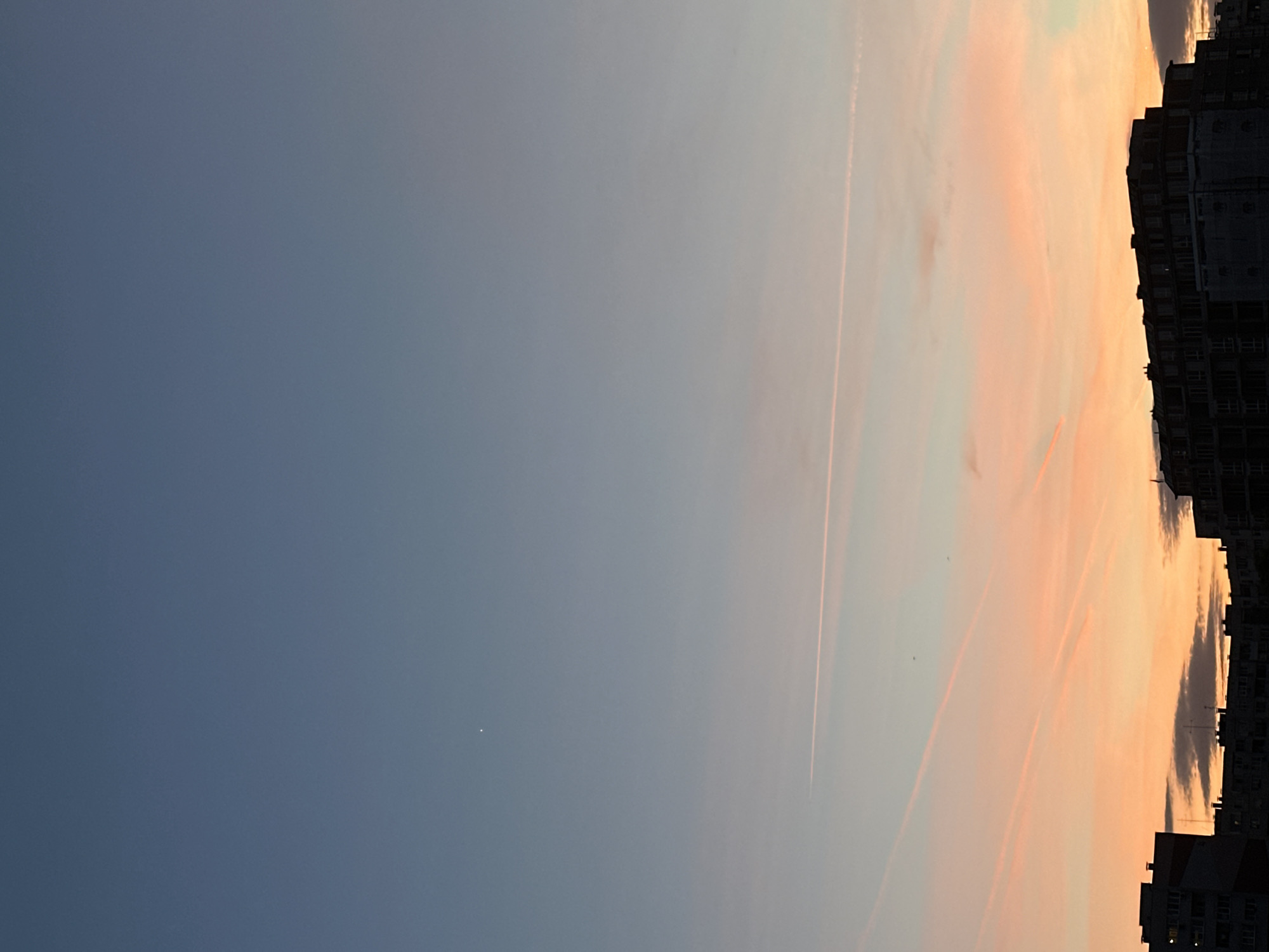 The image shows a sky at sunset over Sofia, Bulgaria. The sky transitions from a deep blue on the left to a pale orange and pink on the right side of the image, where the sun is setting. There are visible contrails from airplanes cutting across the sky. On the right, the silhouette of a building with numerous windows is visible against the colorful sky. A single bright star is visible in the lower left corner of the image.