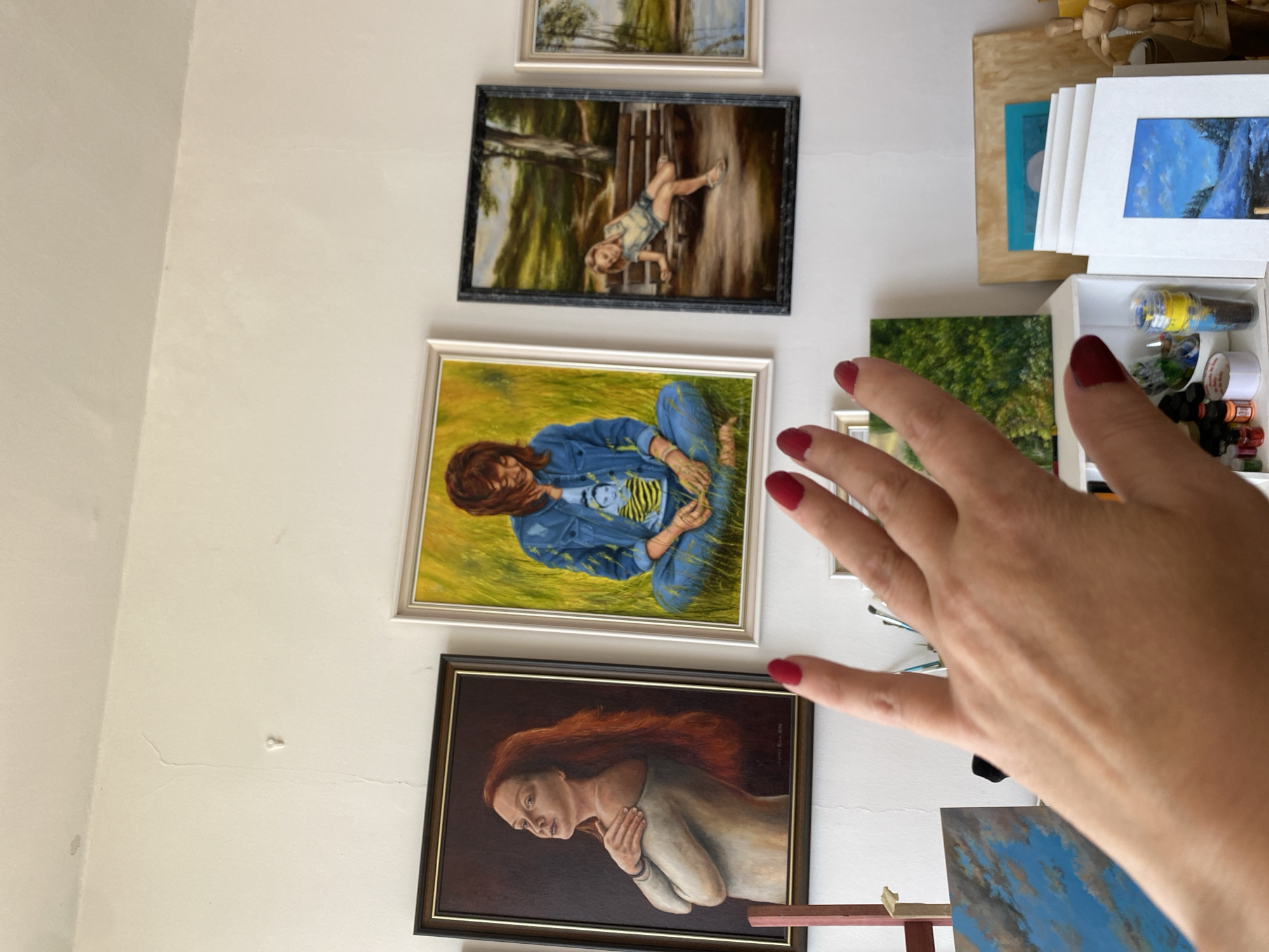 The image depicts an indoor scene, likely an artist's studio or living space, adorned with several framed paintings displayed on a white wall. In the foreground, a woman's left hand, with polished red fingernails, is raised towards the camera, partially obscuring some of the artwork.
The paintings visible are primarily portraits and landscapes. On the far left is a portrait of a woman with long, red hair, looking to the left, her arms crossed over her chest. To its right is a painting of a woman with reddish-brown hair, wearing a denim jacket and blue jeans, sitting cross-legged in a field of yellow grass. Further to the right, a painting shows a young girl in shorts and a light-colored top sitting on a park bench under a tree, with a body of water visible in the distance. Another landscape painting, featuring a body of water and trees, is partially visible on the far right.
Below the wall-mounted artwork, more paintings and art supplies are visible. To the left, a painting of a cloudy blue sky with white clouds is propped up. In the center, a white shelf unit holds various items, including paint tubes, brushes, and a small container with yellow liquid. Further to the right, a collection of matted prints or small paintings are stacked, some showing snowy landscapes. Wooden artist's mannequins are also present on the right side of the frame. The wall itself appears to have some imperfections, including a small crack and a few minor spots.
The overall impression is that of a creative environment, with a focus on the artwork and the artist's presence. The location is identified as Bílina, Czechia.