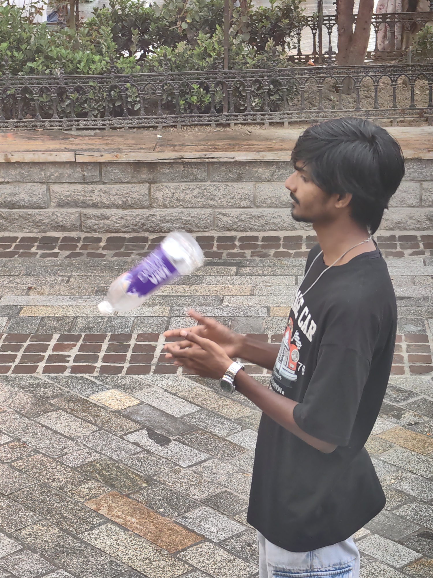 A young South Asian man with dark, tousled hair and a mustache is captured mid-action, attempting to catch a translucent plastic water bottle with a purple label as it tumbles through the air. He is wearing a black t-shirt with a graphic design depicting a vintage vehicle and the number "76", a silver chain necklace, a wristwatch on his left arm, and light-colored denim shorts. His hands are positioned to receive the bottle. The background consists of a textured stone wall and a paved ground made of various colored rectangular stones. Behind the man, a dark metal fence with decorative elements separates the walkway from a green, leafy hedge. The scene suggests an outdoor urban environment, possibly a park or public space in the city of Manali, India.