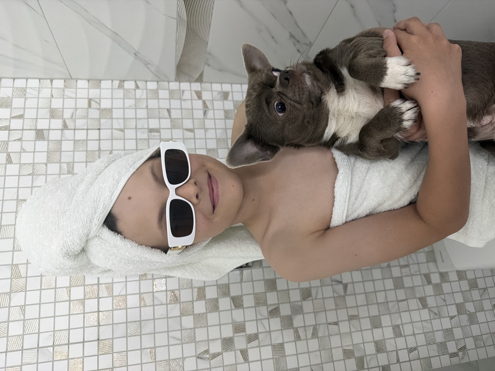 The image shows a child lying on a tiled surface, likely a bathroom floor. The child is wrapped in a white towel and wears white-framed sunglasses. A gray and white puppy rests on the child's chest, interacting with the child's face. The background consists of tiled walls. There is no information to indicate that this image was taken in Votkinsk, Russia.