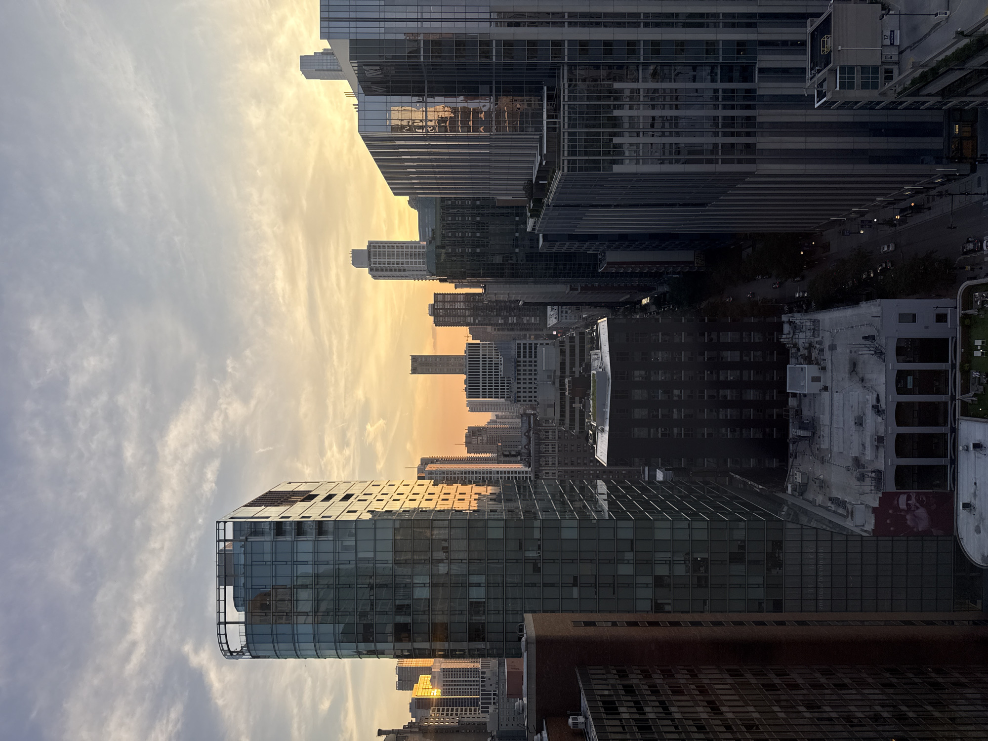An aerial view captures a cityscape at dusk in Chicago, United States of America. The sky is a gradient of soft orange and yellow hues, transitioning to pale blue with wispy clouds. Several tall buildings, constructed with glass and metal facades, dominate the scene, reflecting the sky's colors. In the foreground, a prominent glass-encased building with a curved top stands to the left. To the right, another cluster of modern skyscrapers features reflective surfaces, showcasing vertical architectural elements. Lower buildings and rooftops are visible in the mid-ground and foreground, some with visible rooftop features like HVAC units and green spaces. A street is visible winding between buildings in the distance, with some cars and streetlights. The overall impression is a metropolitan landscape bathed in the warm glow of the setting sun.