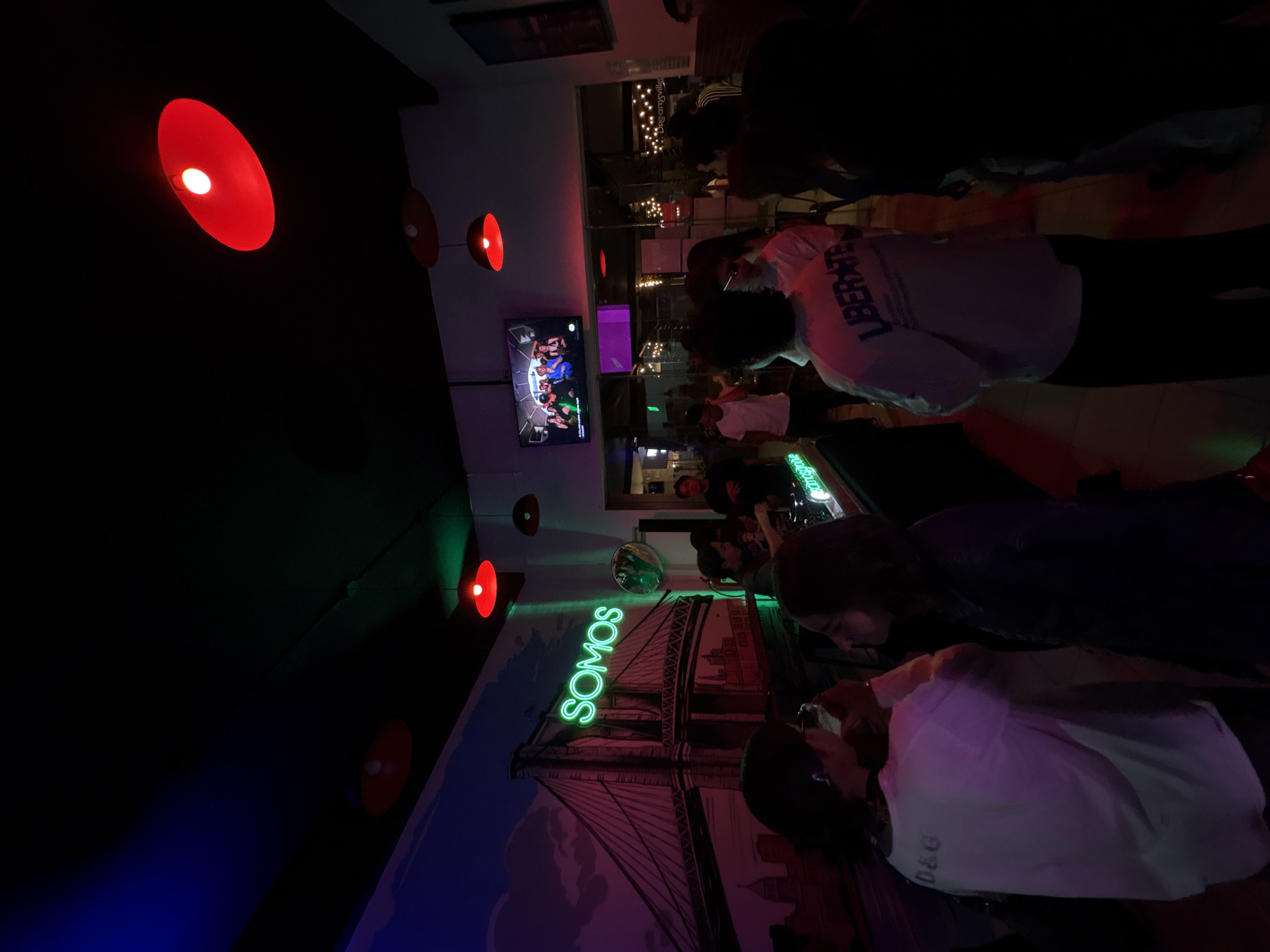 Indoor scene in a bar, likely in San Bernardino Tlaxcalancingo, Mexico. Multiple patrons are present. A mural featuring the word "SOMOS" in green neon lights and a depiction of a bridge (likely the Brooklyn Bridge) is visible on the wall. Red and brown lamps are positioned on the ceiling. A television screen displays an unknown program. People are standing and seated at tables, some engaged in conversation.