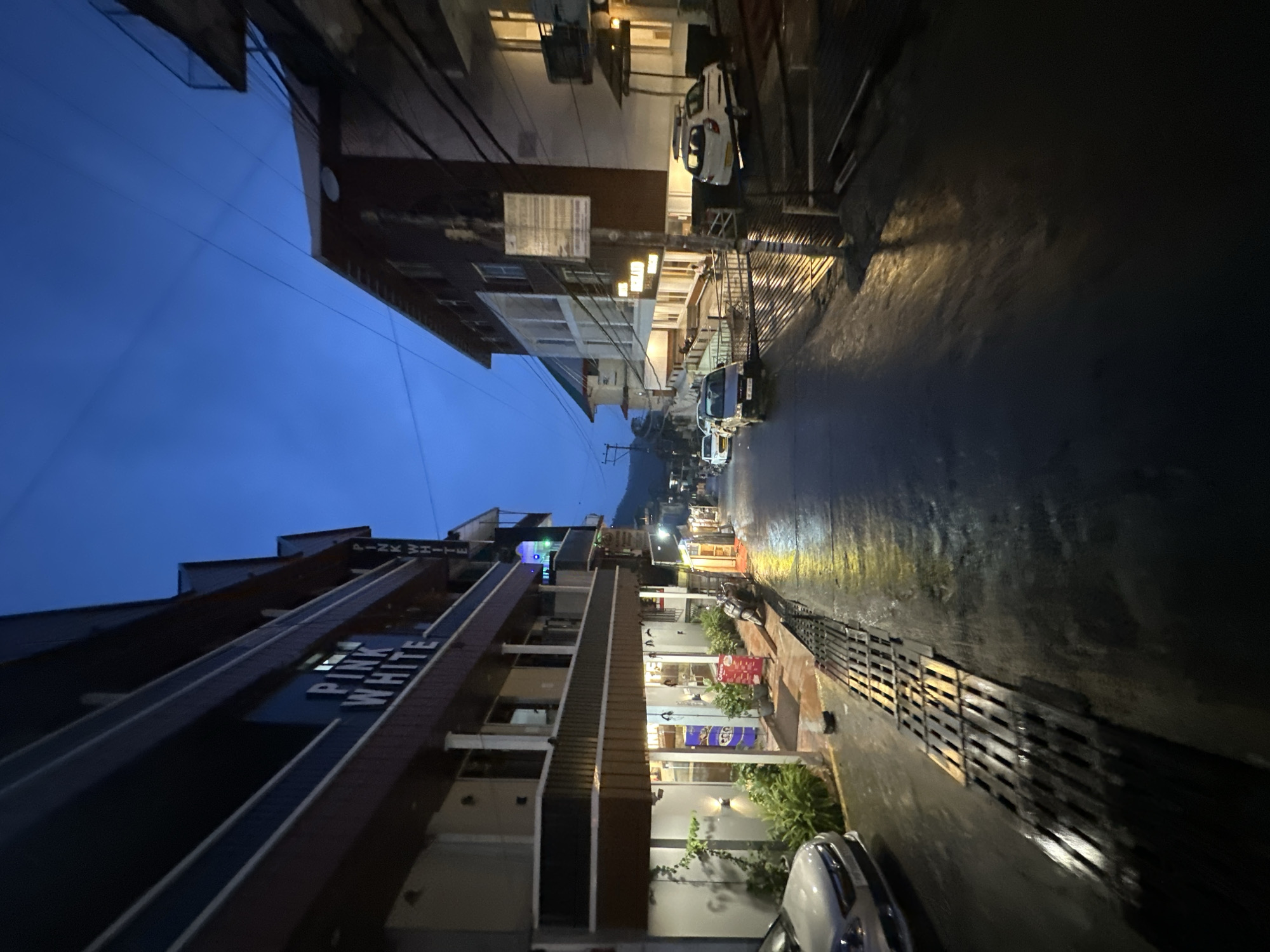 The image depicts a street scene in an urban environment, likely at dusk or in the early evening, given the dark blue sky and artificial lighting. On the left side of the street, a multi-story building with a dark facade and illuminated windows dominates the foreground. A sign on this building reads "PINK WHITE." Below this, the building's lower levels appear to be commercial spaces, with illuminated interiors and plants arranged outside. A silver car is parked on the left edge of the street.
The street itself is wet and reflects the lights from the buildings and street lamps, suggesting recent rain or damp conditions. A metal grate runs along the side of the street in the foreground. Several cars are parked and driving along the street, which recedes into the distance. On the right side of the street, another multi-story building is visible, also with illuminated windows. The overall atmosphere is one of a quiet, possibly small-town or city street at the end of the day.