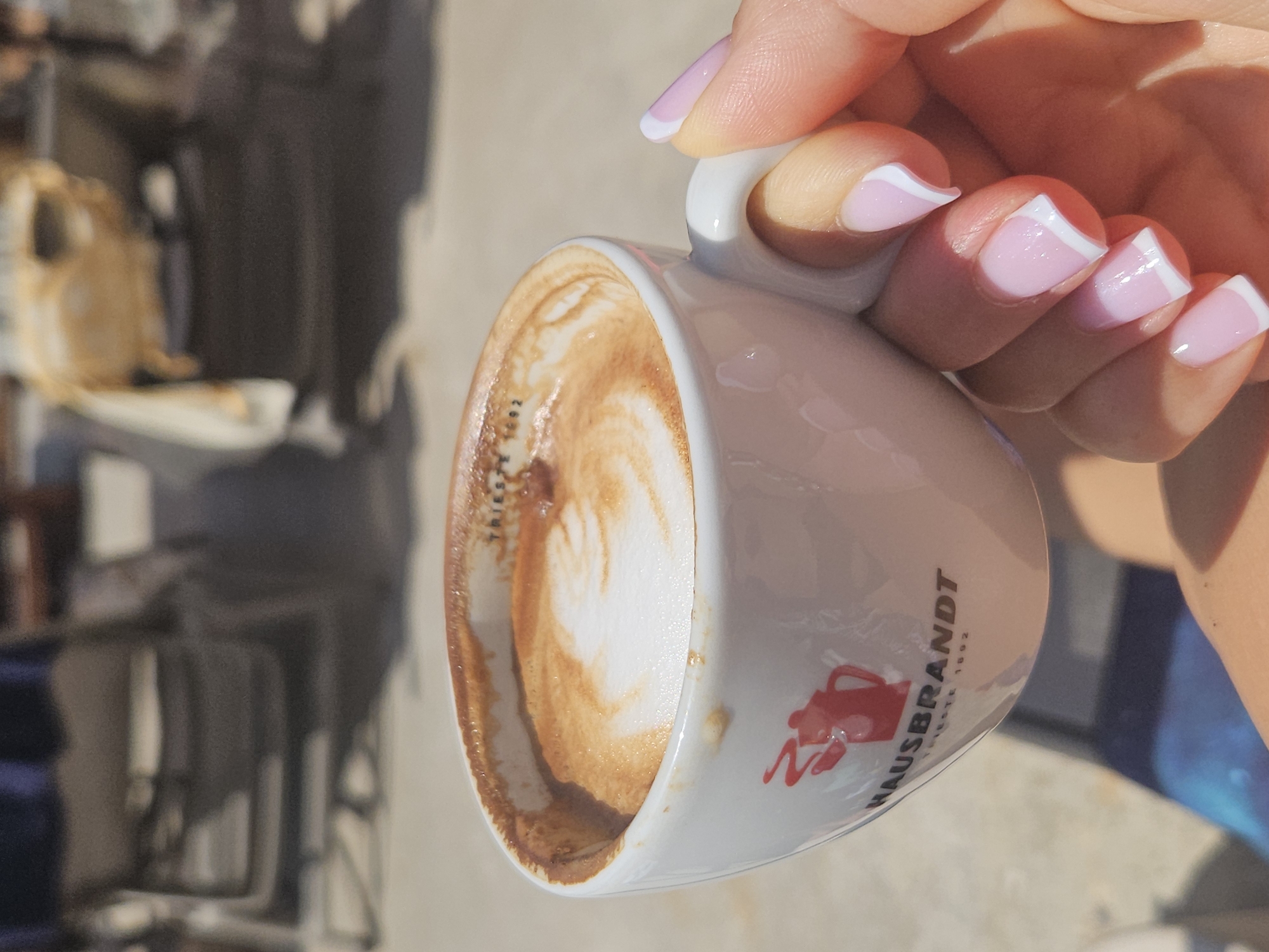 A close-up shot shows a hand with pink and white French manicured nails holding a white coffee cup with a Hausbrandt logo. The coffee in the cup has foam art. The background is out of focus and appears to be an outdoor seating area with chairs.