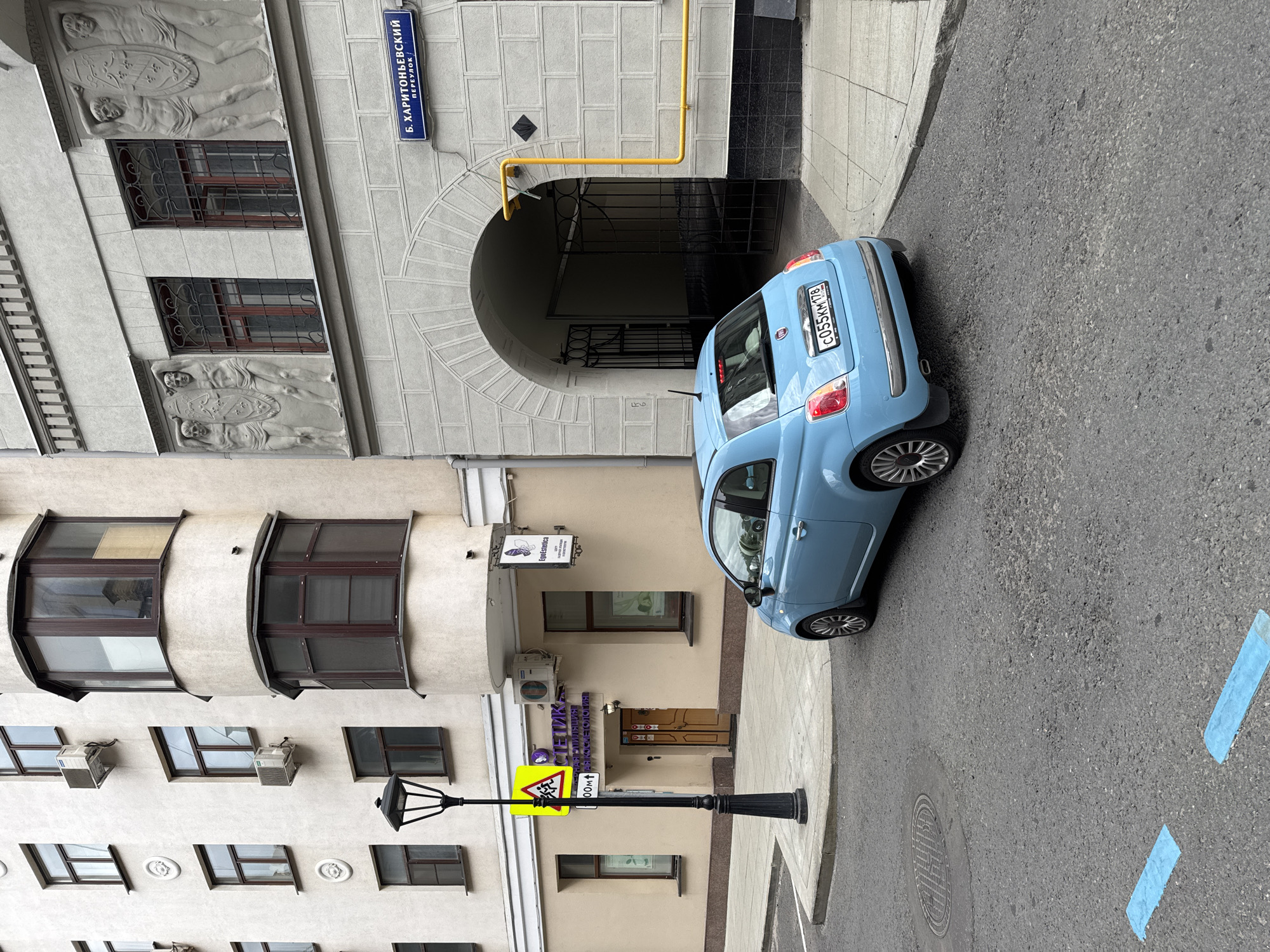 The image depicts a street scene in Moscow, Russia, specifically on Kharitonievskiy Lane. The focal point is a light blue Fiat 500 parked on the asphalt road. To the left, a cream-colored building with multiple windows and air conditioning units is visible. A lamppost stands on the sidewalk in front of this building, next to a yellow warning sign depicting children crossing. Further to the left, a doorway with a wooden door is partially visible.
To the right of the Fiat, a light gray building with an arched entranceway is featured. Above the entrance, a street sign reads "Б. ХАРИТОНЬЕВСКИЙ / ПЕРЕУЛОК" (B. Kharitonievskiy Lane). Sculptural figures are present on the facade of this building, above two windows. A yellow gas pipe runs vertically along the right side of the gray building.
The road surface is asphalt, with two blue markings visible in the foreground. The overall environment suggests an urban setting with residential or commercial buildings lining the street. No people are clearly visible in the image.