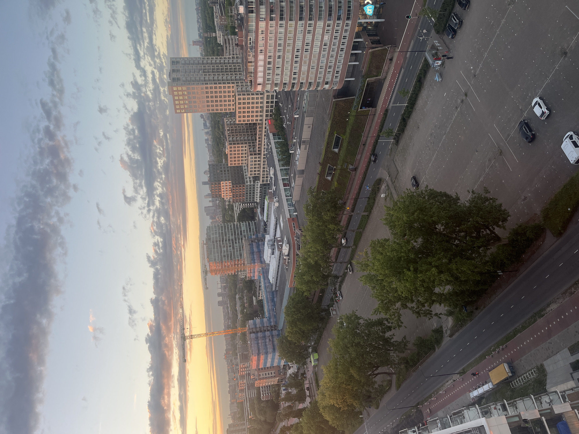 An aerial view captures a city skyline at dusk with a warm, orange glow from the setting sun. Several tall buildings, some modern and glass-fronted, others older and brick, punctuate the horizon. Construction cranes are visible, indicating ongoing development. A tree-lined street with a bike path runs through the scene, with cars parked in a large lot and driving on the road. Some people are visible walking near a grocery store. The sky is a mix of pale blue and scattered clouds, tinged with pink and orange from the sunset.