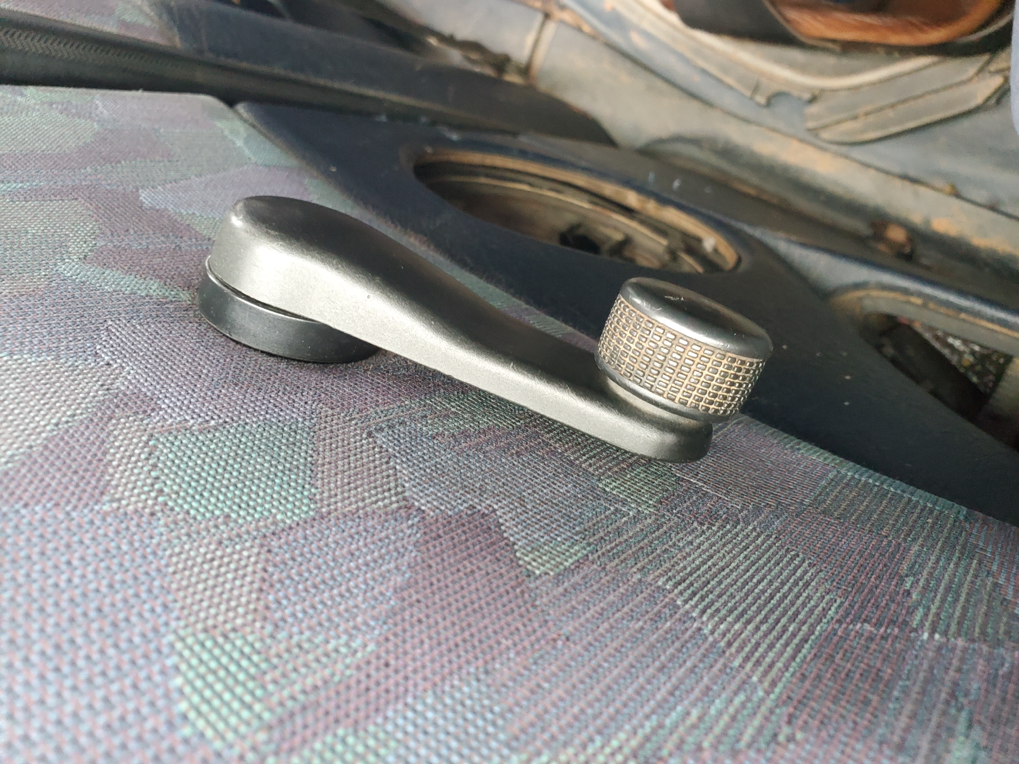 This is a close-up, eye-level, medium shot of a car's interior door handle and part of the door panel. The door panel is covered in a patterned fabric with shades of purple, green, and gray. A silver-colored window crank with a textured grip is mounted on the door panel, positioned horizontally. To the right of the crank, a dark-colored opening is visible, possibly where a speaker or other car component would be. The bottom right of the image shows a glimpse of a person's brown shoe and part of what appears to be a seat or floor mat. The lighting suggests an indoor or shaded environment. The scene is not located in Pankshin, Nigeria.