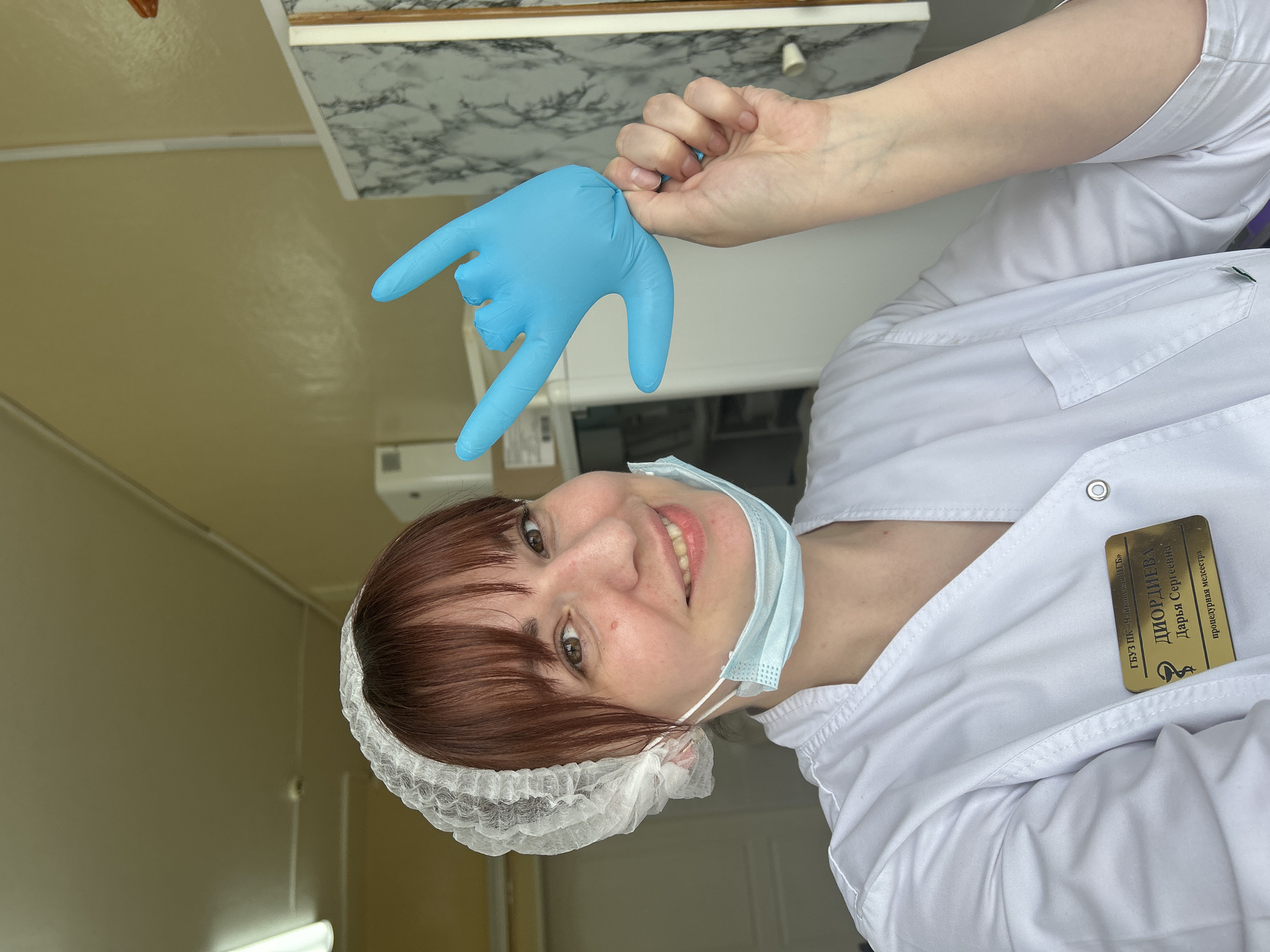 A smiling woman in medical attire is captured in a selfie-style portrait within what appears to be an indoor clinical or medical setting in Fok, Russia. She has brown hair with bangs, brown eyes, and is wearing a white medical coat, a white disposable hair net, and a light blue surgical mask pulled down to her chin. Her left hand, adorned with a bright blue medical glove, is raised, forming a hand gesture with the index finger, pinky, and thumb extended, often associated with "rock and roll" or "I love you." The woman looks directly at the camera with a cheerful expression.
The background shows light-colored walls, possibly beige or light yellow, and a white ceiling with visible fluorescent lighting fixtures. A white shelf or counter with a grey marble pattern is also visible in the upper right. On the left side of her chest, a gold-colored name badge is pinned to her white coat. The text on the badge is clearly visible and reads:
"ГБУЗПК «Чайковская ЦРБ»"
"ДИОРДИЕВА"
"Дарья Сергеевна"
"процедурная медсестра"
This translates to "State Budgetary Healthcare Institution of Perm Krai 'Tchaikovsky Central District Hospital'", "DIORDIEVA", "Darya Sergeevna", and "Procedure Nurse". The overall scene suggests a professional yet friendly atmosphere, likely within a healthcare facility.