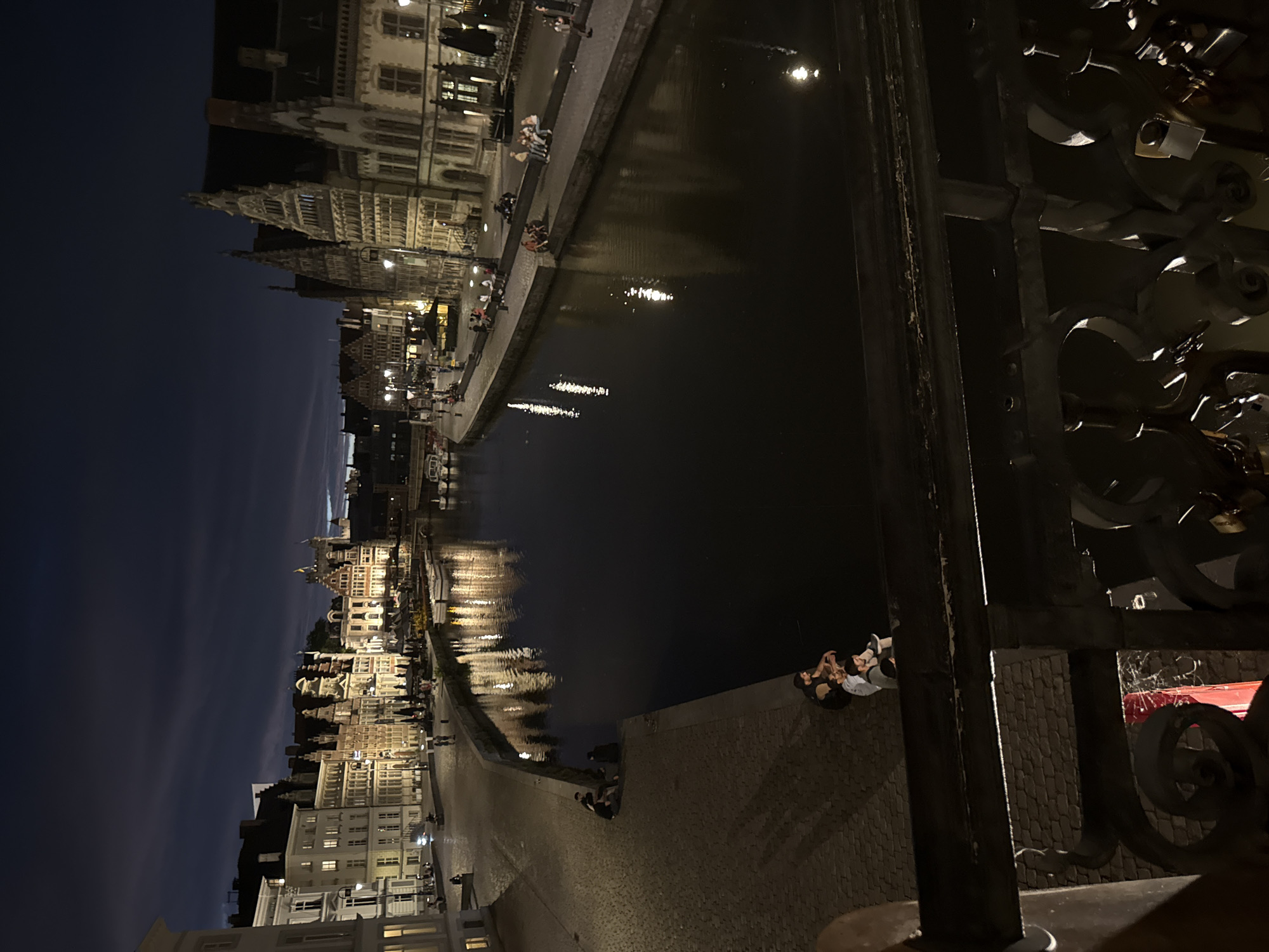 This is a nighttime, long-exposure photograph taken from a bridge overlooking a canal in Ghent, Belgium. The canal runs horizontally across the frame, reflecting the lights of the historic buildings lining its banks. On the left side of the canal, a cobblestone promenade is visible, with several groups of people sitting or walking along it. The buildings are characteristic of Ghent's architecture, with gabled roofs and illuminated windows. On the right side of the canal, more buildings are visible, also lit up, and a few boats are moored. The sky above is dark blue with streaks of clouds, and the water is dark, with shimmering reflections of the lights. The foreground of the image shows the dark, ornate railing of the bridge from which the photo was taken, with several padlocks attached to it, suggesting it is a "love lock" bridge. In the lower-left corner, a bright red object is partially visible. The overall atmosphere is tranquil and picturesque, capturing the beauty of Ghent at night.