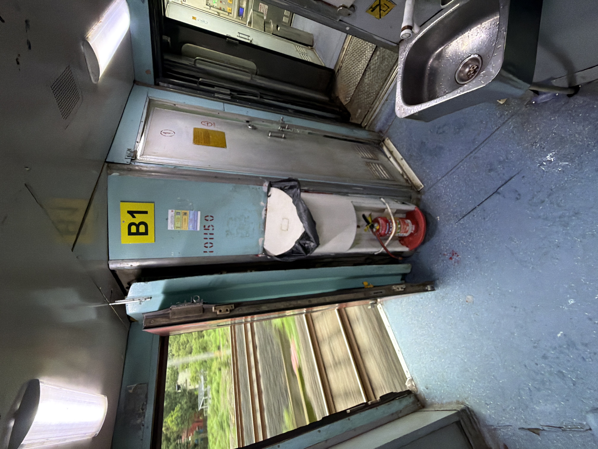 The image depicts the interior of a train carriage, likely a passenger compartment in Saphale, India. The view is from inside the carriage, looking towards the open doorway and a corridor. The floor is a light blue, speckled with some debris and what appears to be water or liquid stains. To the left, an open doorway reveals train tracks and blurred greenery outside, indicating the train is in motion.
Inside the carriage, a teal-colored wall features a yellow sign with the letter "B1". Below this, a number "101150" is stenciled in red. Adjacent to this is a white, wall-mounted water dispenser with a black bag covering the top of the container. A red fire extinguisher is visible at the base of the dispenser.
To the right of the dispenser is a silver door with a circular symbol and a yellow notice. Further into the carriage, another silver door is visible, partially open, leading to a different section. In the foreground on the right, a stainless steel sink with a faucet is positioned. The sink shows signs of recent use with water droplets. The overall environment suggests a functional, albeit somewhat worn, train interior. There are no people visible in the frame.