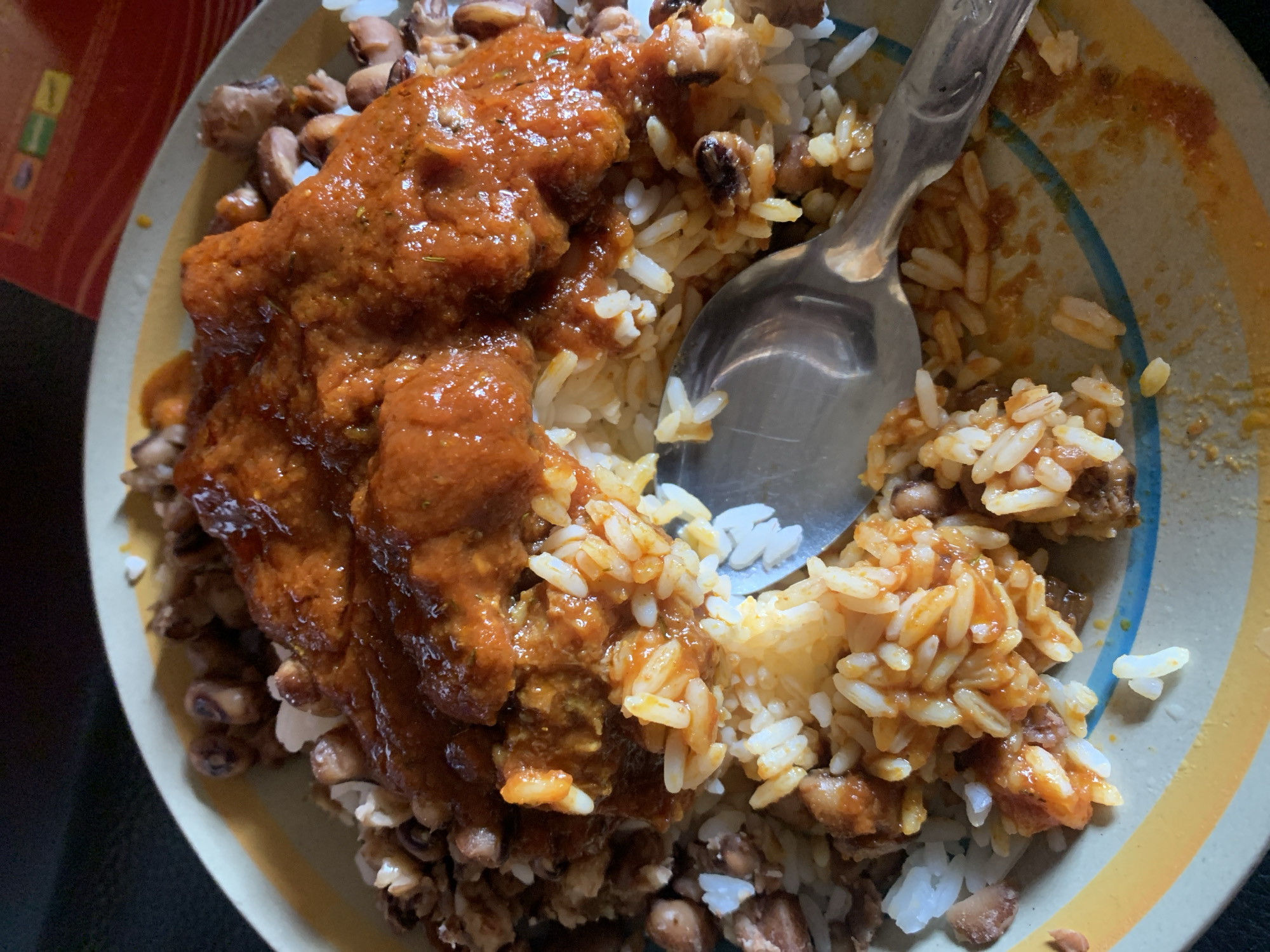 A plate of food containing rice, beans, and stew is visible. A silver spoon rests on the rice. The plate has a multi-colored rim. The food is positioned in the center of the plate. The background includes a partial view of what appears to be a container, along with a dark surface. The location is Rafin Par, Nigeria.