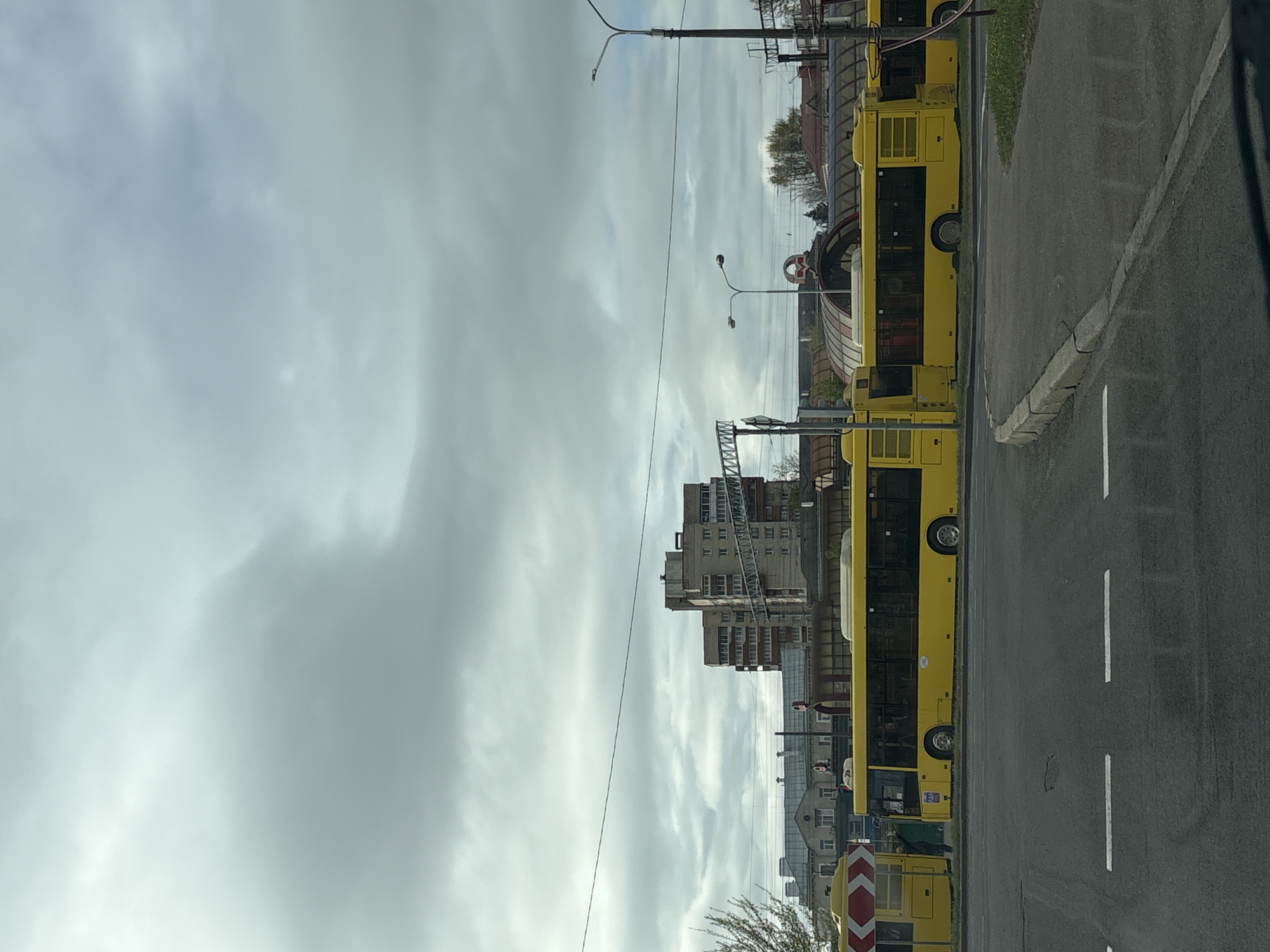 The image depicts a street scene in Minsk, Belarus, with yellow buses parked along the right side of the road. The road has white dashed lane markings. Buildings, including a high-rise apartment block, line the opposite side of the street. Overhead power lines are visible against a cloudy sky. A "M" symbol, often associated with the Minsk Metro, is visible in the background. A chevron traffic sign is visible on the lower right.
