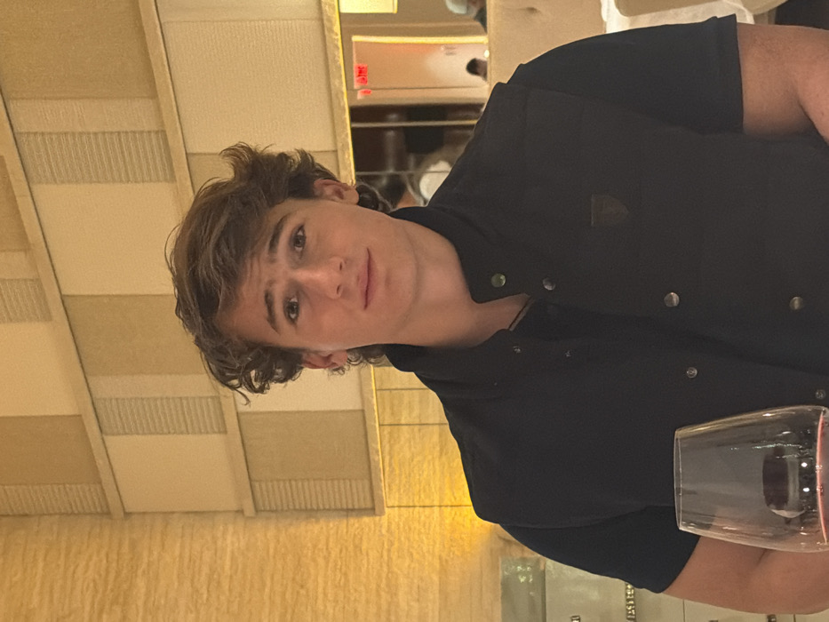 A young man with curly brown hair looks at the viewer. He wears a dark blue button-up shirt. The background includes a textured wall and part of a wine glass. The image appears to be taken indoors, possibly in a restaurant, judging by the ambient lighting and decor.