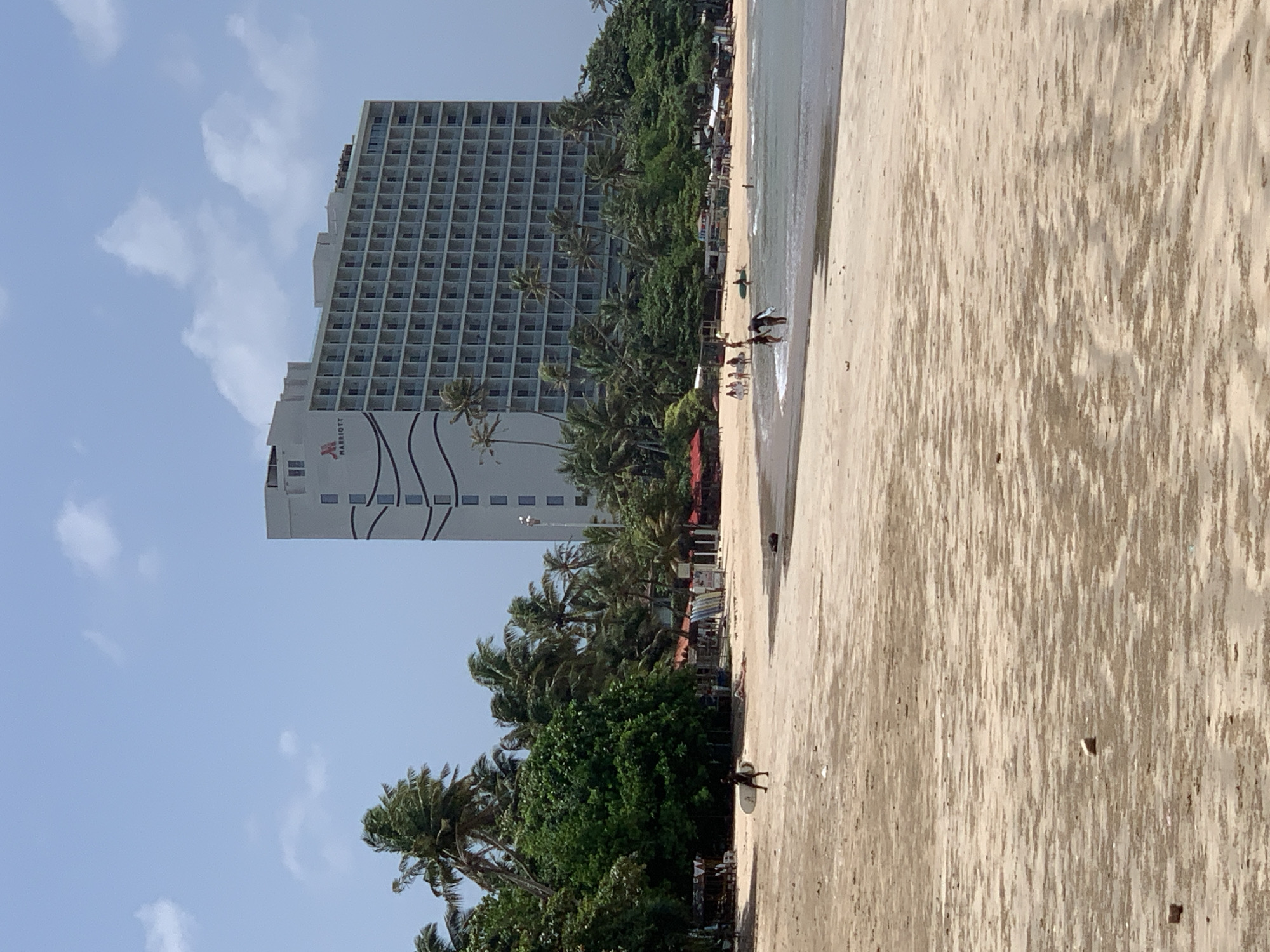 The image depicts the Weligama beach in Sri Lanka, featuring the iconic Marriott hotel in the background. The foreground is dominated by the sandy beach with a few scattered debris. In the mid-ground, several people are seen walking along the shoreline and in the shallow water, some carrying surfboards. The beach is lined with palm trees and dense green foliage, with small stalls or shops visible among the trees. The ocean is calm, with gentle waves lapping at the shore under a partly cloudy sky.
