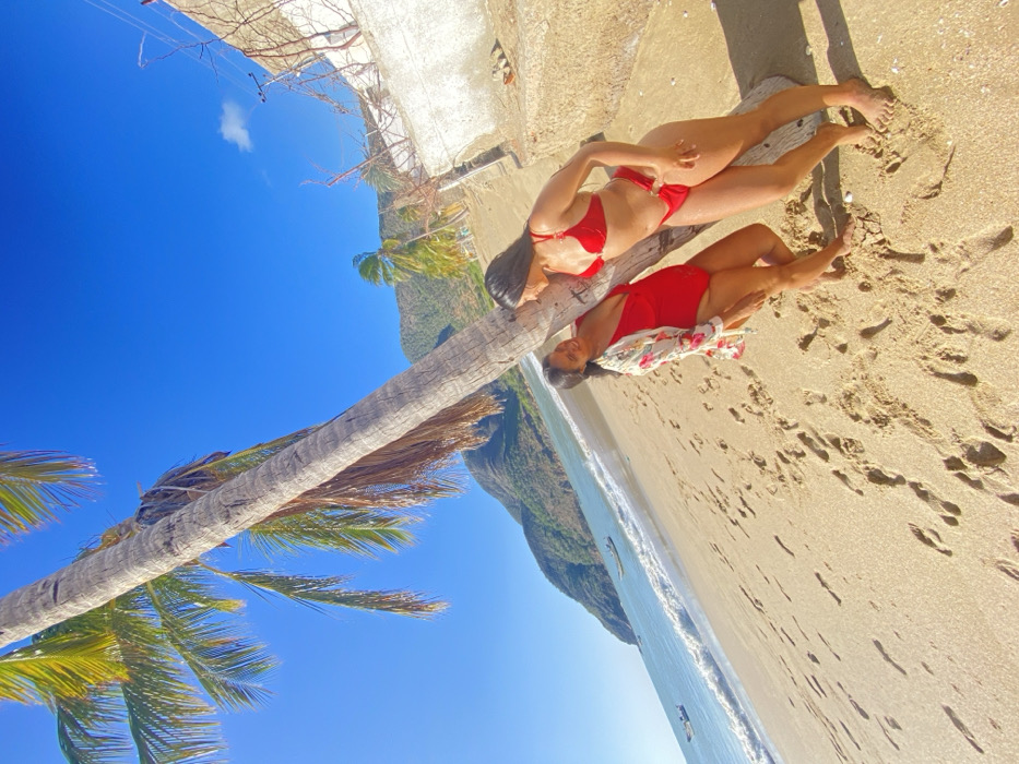 Two women in red swimwear pose by a fallen palm tree on a sandy beach. One wears a bikini, the other a one-piece swimsuit with a floral cover-up. The turquoise ocean and a mountain are visible in the background. Clear blue sky and palm fronds frame the scene. The ground is covered with footprints in the sand. The orientation of the photo is rotated so that the sky is angled downwards.