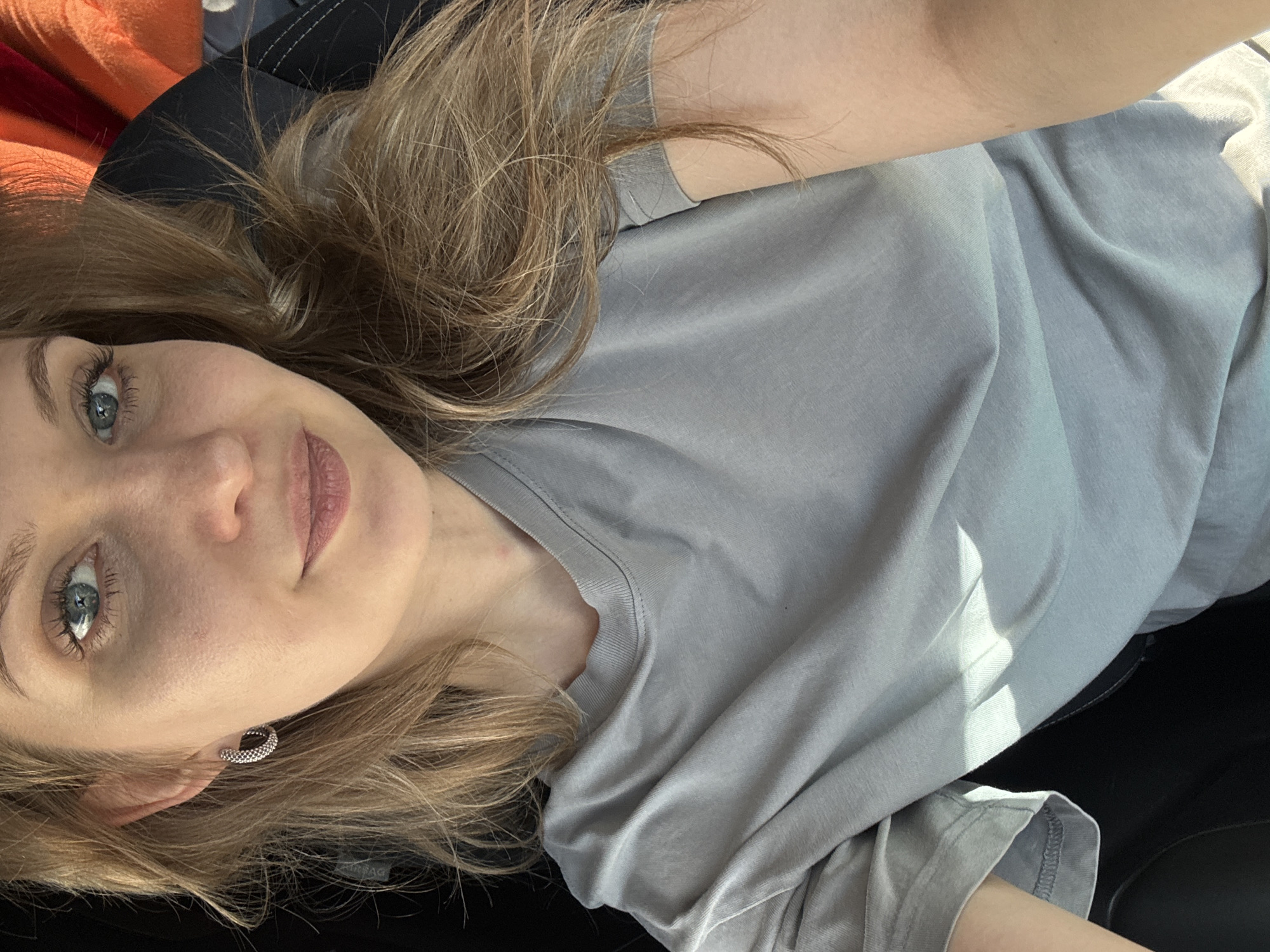 A woman is lying down, wearing a light gray t-shirt and small hoop earrings. Her light brown hair is styled with loose waves. The setting appears to be inside a car, based on the dark interior. The location, Izhevsk, Russia, cannot be determined from this image alone.