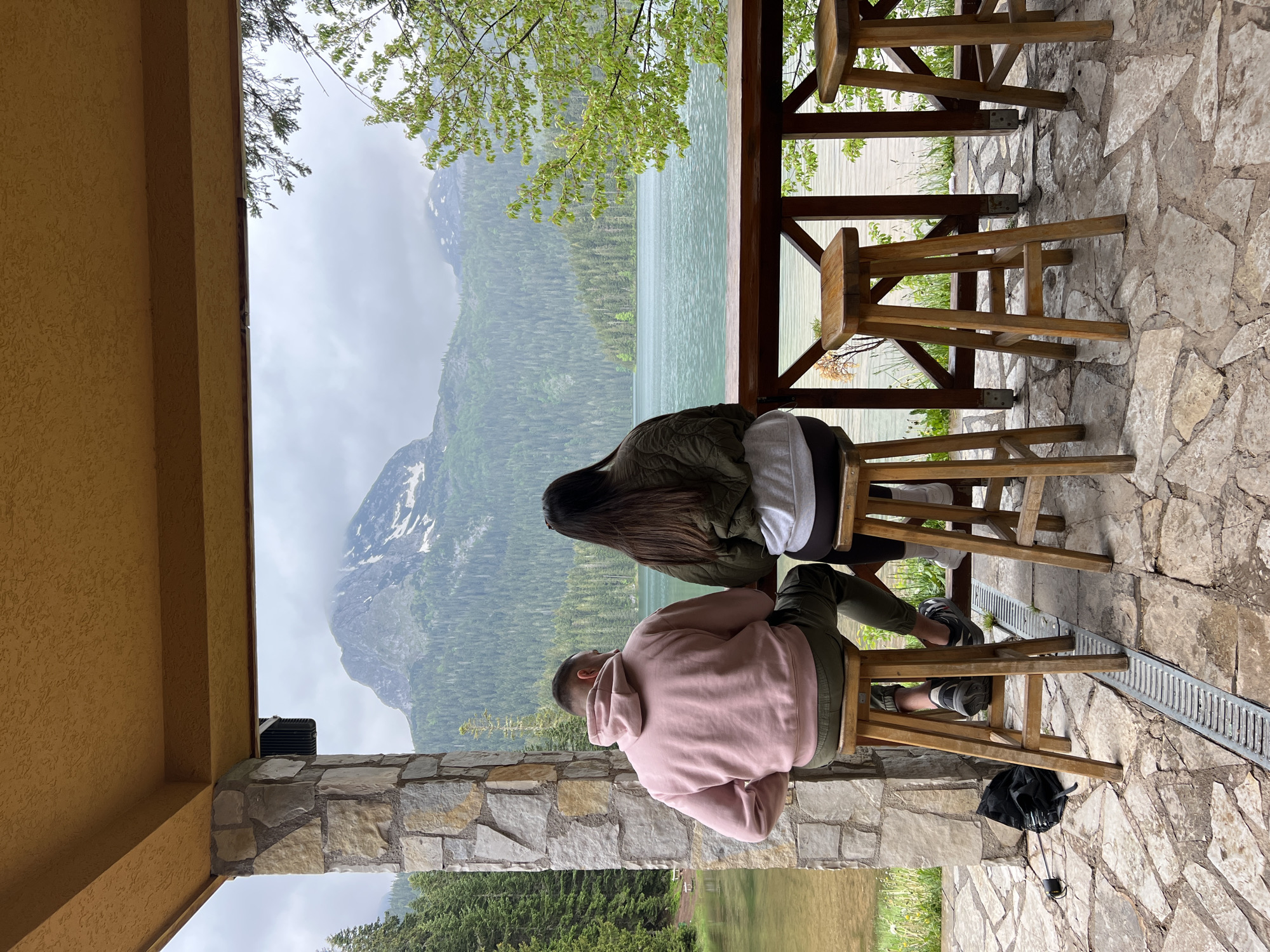 This image captures a serene outdoor scene from a sheltered viewpoint, likely a cafe or observation deck, overlooking a magnificent natural landscape. Two individuals are seated on high wooden bar stools, their backs to the camera, engrossed in the view.
The person on the left is wearing a pink hoodie and olive green pants, with short dark hair. The person on the right has long dark hair and is dressed in a dark green jacket over a white top and dark leggings. They are looking out towards a large, light green-blue lake, which is bordered by dense green forests. Beyond the forests, rugged mountains with visible patches of snow rise under an overcast sky.
The immediate foreground features a textured stone floor made of irregular flagstones. A sturdy wooden railing system separates the seating area from the expansive view. To the left, a stone wall or pillar is visible, capped by a yellow-stuccoed overhang or ceiling. Sparse green tree branches hang into the frame from above. On the floor near the stools, a black bag and a metal drainage grate can be seen.
The atmosphere is peaceful and contemplative, with the cloudy weather adding a soft, diffused light to the dramatic mountain and lake scenery. The time of day is daytime, though the overcast conditions prevent bright sunshine. No visible text is present in the image. This picturesque setting is located in Pitomine, Montenegro.
