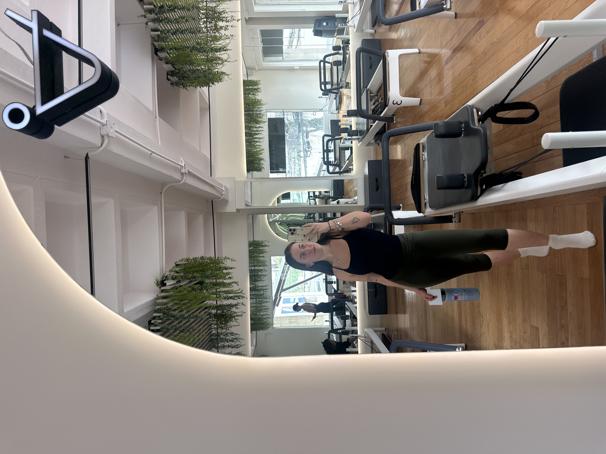 A young woman stands in a brightly lit, modern Pilates studio. The studio is located in Jesús del Monte, Mexico. The studio features wooden floors, white walls, and large windows. The woman is positioned in front of a mirror, using her cell phone to take a selfie. She is wearing a black tank top, olive-green leggings, and white socks. She is holding a water bottle in her left hand. Reflected in the mirror are other Pilates machines and people exercising. The studio is decorated with greenery in white shelving units. A distinctive sign is visible outside the studio's entrance.