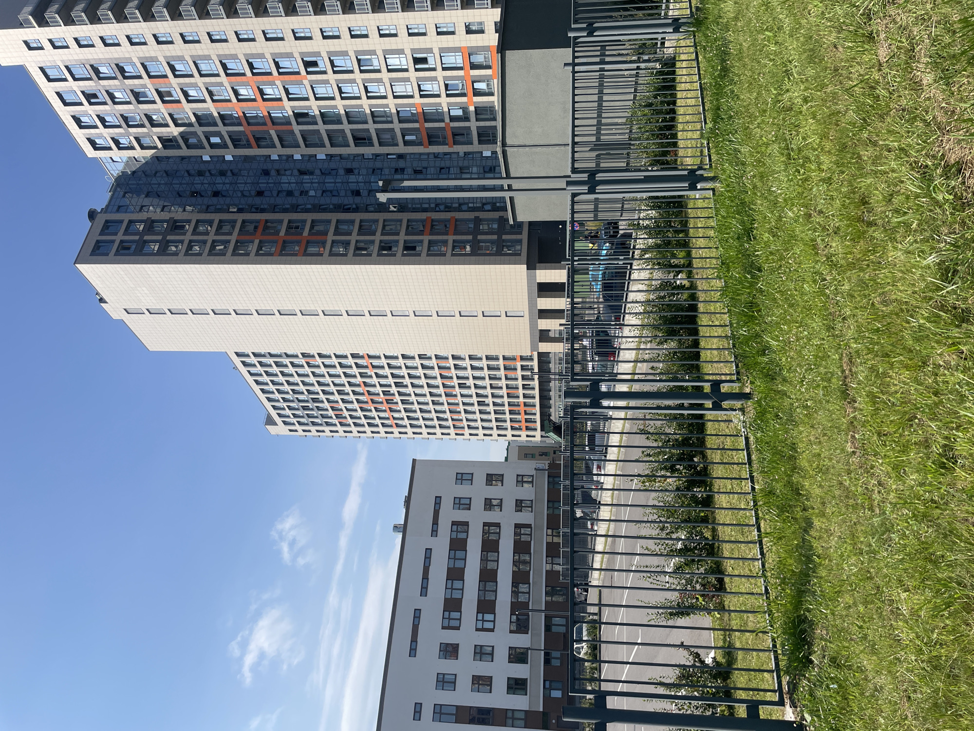 The image shows a view of a residential complex in Pargolovo, Russia. In the foreground, there is a metal fence and a patch of green grass. Behind the fence, there is a paved parking lot with several cars visible. Further in the background, there are modern apartment buildings with a contemporary architectural style, characterized by large windows and a combination of light-colored facades and orange accents. The sky is blue with a few wispy clouds.