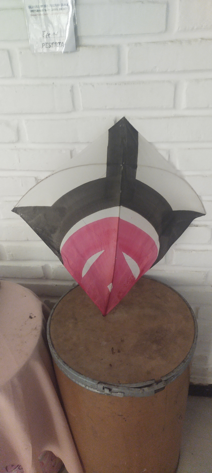 A colorful kite is displayed in front of a white brick wall. The kite has a diamond shape with black and white patterns, and a pink section with two white crescent shapes. It rests on top of a round, wooden object, possibly a drum or a barrel, which has a metal band around its rim. To the left of the kite, a portion of a pink fabric-covered object, likely a table or chair, is visible. A laminated paper with Indonesian text is attached to the brick wall above the kite. The environment appears to be indoors.