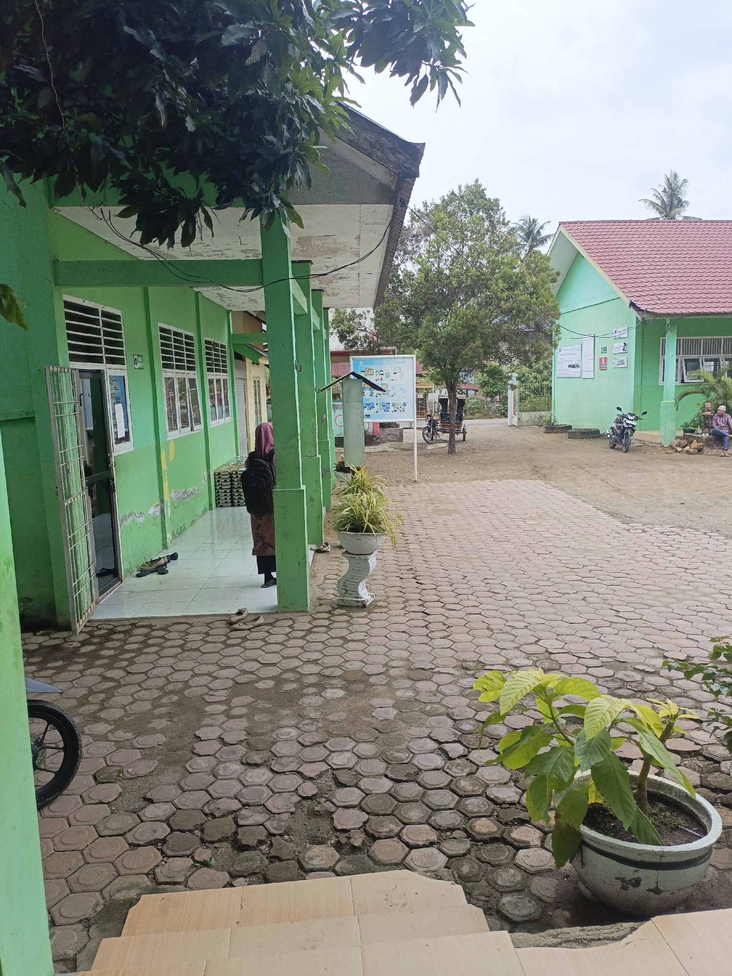 The image depicts an outdoor scene at what appears to be a school or public building in Lhokseumawe, Indonesia. The primary structure is painted a vibrant green and features a covered walkway with supporting pillars. Several windows with horizontal louvers are visible along the building's facade. In the foreground, a woman wearing a pink hijab and a brown and black patterned skirt stands with her back to the camera, carrying a backpack. Footwear, including sandals, is scattered on the tiled walkway.
The ground is paved with interlocking hexagonal concrete blocks, some of which are uneven and surrounded by soil. A potted plant with large green leaves sits in the lower right portion of the frame. In the background, another green building with a red tiled roof is visible, as are trees and palm trees. A motorcycle is parked on the right side of the scene, and a three-wheeled vehicle (likely an auto-rickshaw) is positioned further back in the courtyard. Posters or notice boards are affixed to the wall of the building on the right. The sky is overcast, suggesting a cloudy day. The overall impression is of a utilitarian and functional outdoor space within an Indonesian urban or suburban setting.