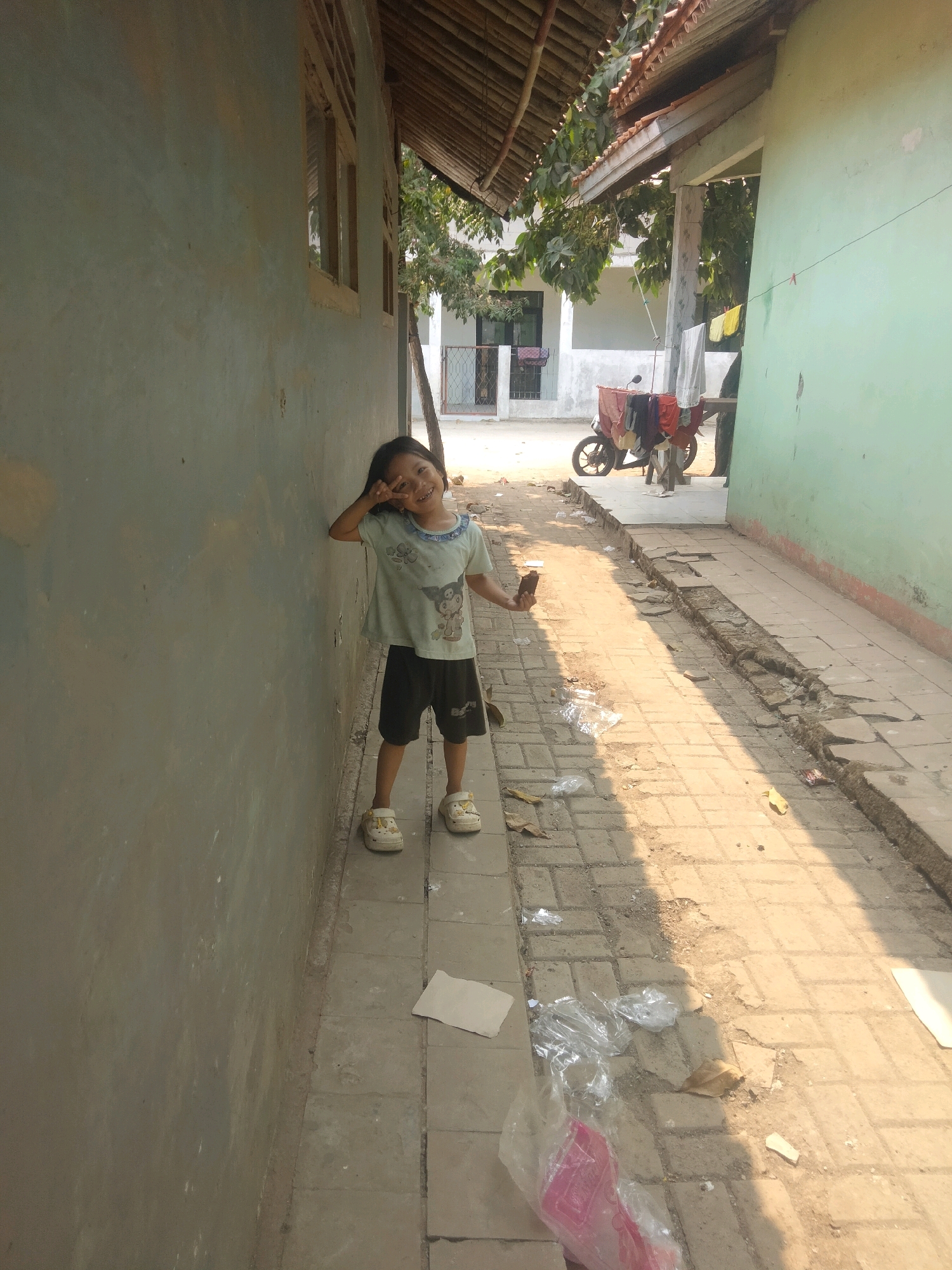 In the city of Suka Sari, Indonesia, a young girl stands on a paved walkway next to a weathered, light-blue wall. She is wearing a light-colored t-shirt with a cartoon graphic and dark shorts, along with light-colored clogs. The girl is smiling and holding her right hand up, forming a peace sign. To her right, the walkway appears damaged with sections missing and debris scattered on the ground, including plastic bags and leaves. Beyond the walkway, a motorcycle is parked with laundry draped over it. The background shows residential buildings with wooden roofs and trees. The scene is illuminated by sunlight, casting shadows on the walkway.