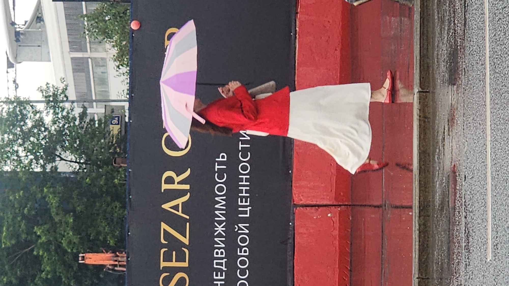 A full-shot, eye-level, outdoor photograph captures a woman walking under a pastel-striped umbrella on a wet sidewalk in Moscow, Russia. The woman, seen from the side, is wearing a red jacket and a long, flowing white skirt. Her feet are clad in red ballet flats, and she carries a beige bag over her shoulder. The umbrella is predominantly pink and purple. The background features a large black banner with gold lettering, which reads "SEZAR GRUPP" in Cyrillic, followed by "НЕДВИЖИМОСТЬ ОСОБОЙ ЦЕННОСТИ". Behind the banner, a blurred industrial crane is visible. The ground is wet and reflective, indicating recent rain. The scene is bordered by bright red concrete barriers in the foreground.