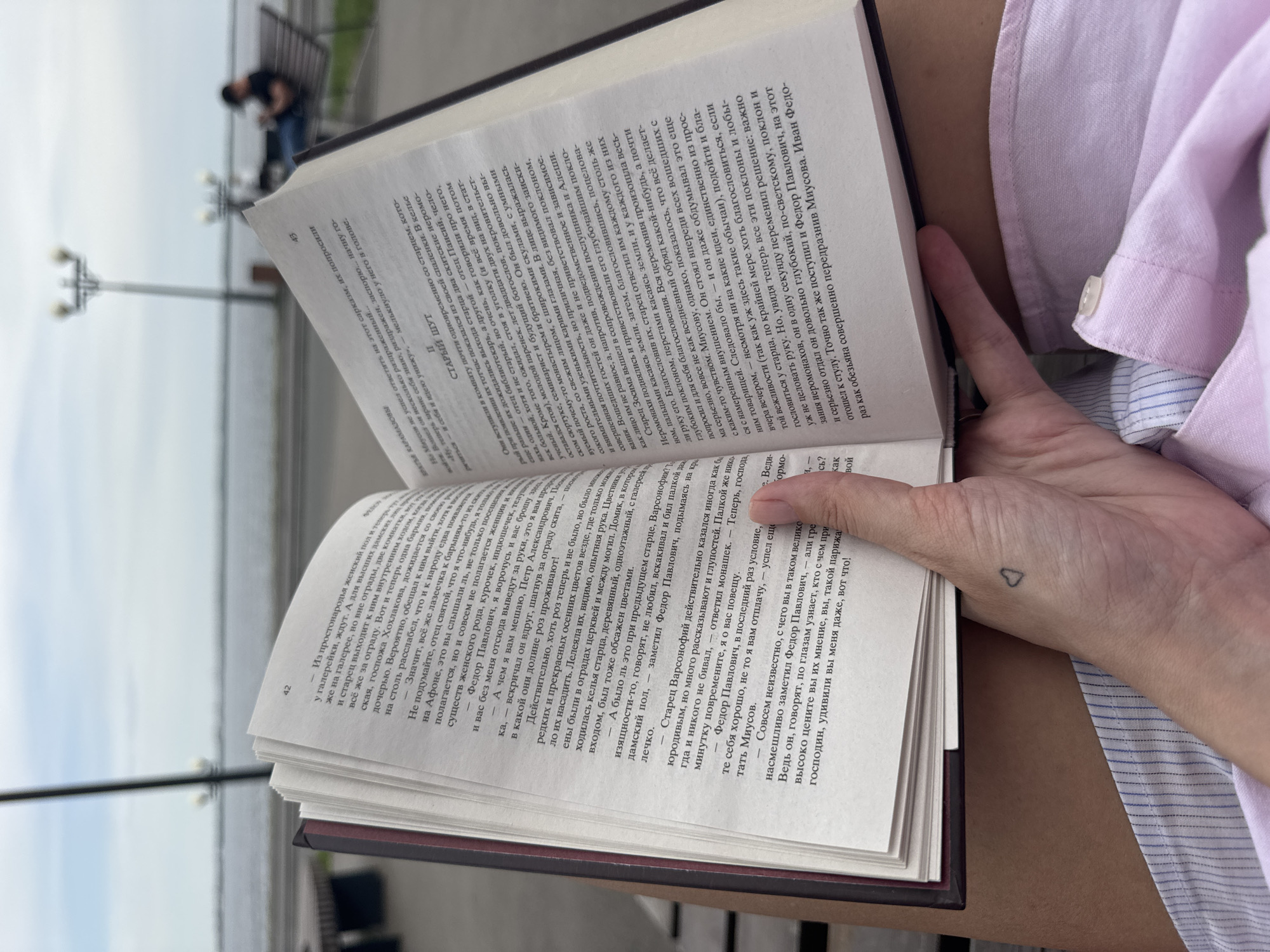 Here's a detailed description of the image:
**General Overview:** The image shows a person holding a book open, presumably reading it. The setting appears to be outdoors, possibly near a waterfront, in Saratov, Russia. The focus is on the book and the reader's hand, with some of the background visible.
**People:** The primary visible person is only partially shown, their hand and forearm are visible. The person is wearing light-colored clothing (possibly a pink shirt) and has a small heart tattoo on their wrist.
**Subjects & Objects:**
* **Book:** The book is open and being held by the person. The pages are filled with text in Cyrillic alphabet, which is likely Russian. The book's spine is visible, appearing dark in color.
* **Background (Outdoors):** There is a waterfront area with benches, lampposts, and other people in the background. The distant figures are blurred, suggesting they are out of focus. The city has modern architectural elements.
* **Clothing:** The clothing of the person holding the book is visible: likely a light pink shirt.
* **Tattoo:** A small, minimalist heart tattoo is present on the person's wrist.
**Actions & Interactions:** The primary action is someone reading a book. The person is holding the book open, indicating they are actively engaged in reading.
**Environment:**
* **Location:** The image is near a waterfront, potentially a river or large body of water. It's likely taken in Saratov, Russia, based on the setting and architectural style.
* **Weather/Time:** The daylight suggests it is daytime. The weather is likely pleasant, as someone is comfortable sitting and reading outside.
**Additional Details (From Text on the Book Pages):** The text appears to be literary in nature. There are mentions of "отец" (father) and "господин" (mister), suggesting a story with characters and dialogue. The text on the left page mentions characters and elements like church, old lady and cemetery, while the text on the right page involves names like "Миусов" and "Федор Павлович" as well as legal terms/agreements, implying that the book is either a novel, a biography, or other work of literature. The pages shown appear to be pages 42 and 43 of the book.