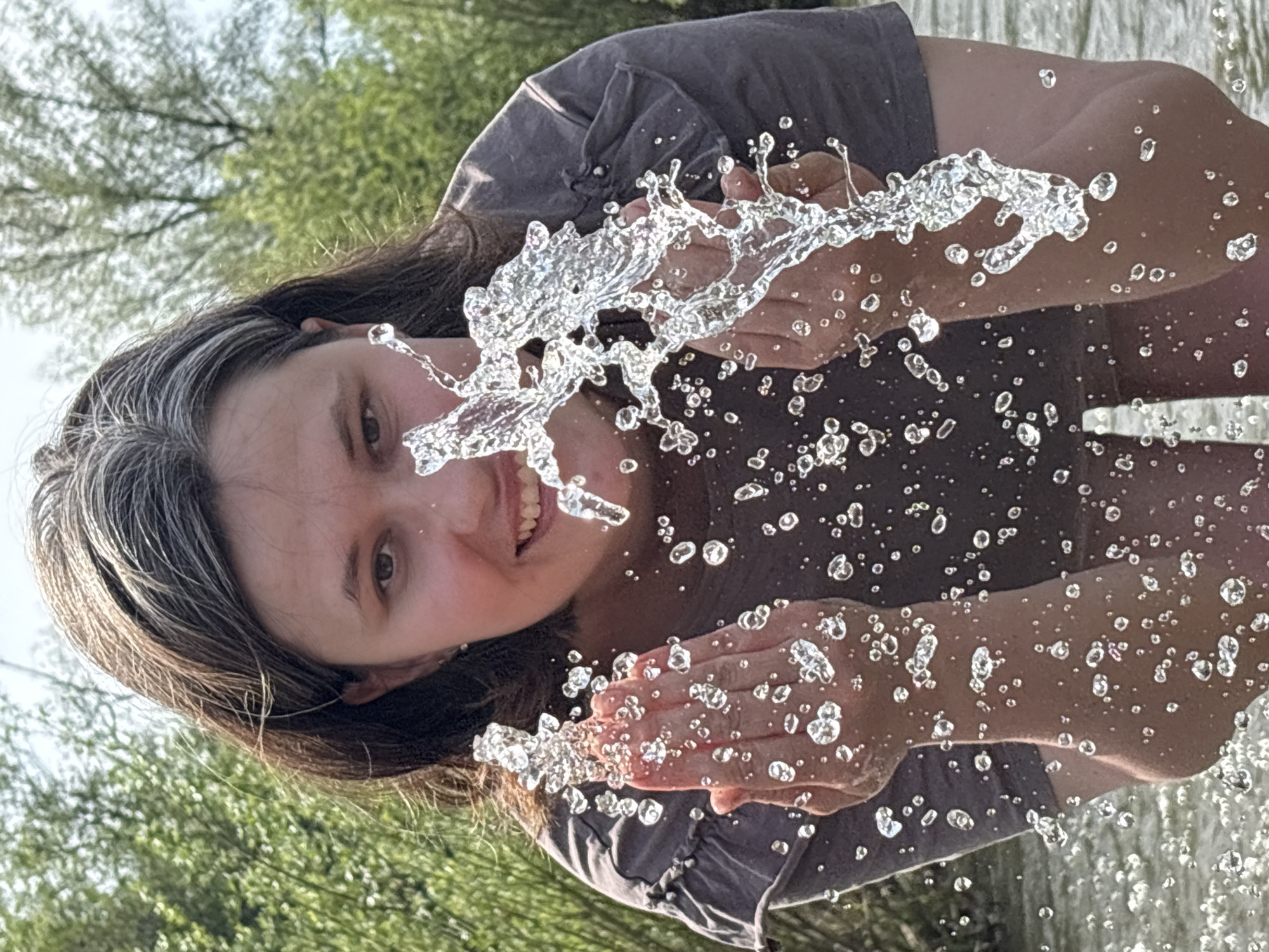 A woman is splashing water in front of her face. She is wearing a brown t-shirt and has brown hair with grey streaks. The background is a blurry green, suggesting trees and foliage. The location, Nizhnyaya Chermoda, Russia, cannot be confirmed solely from visual information.