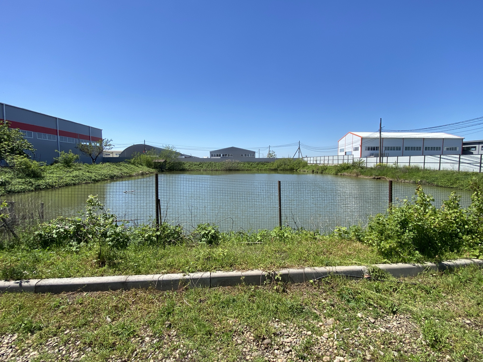 A low-angle daytime shot of a pond in Neftyanik, Russia, bordered by a chain-link fence and vegetation. In the background, several industrial buildings are visible under a clear blue sky. The foreground includes a concrete curb, grass, and small rocks. The pond water is a murky grey-green color. There are no people visible in the image.