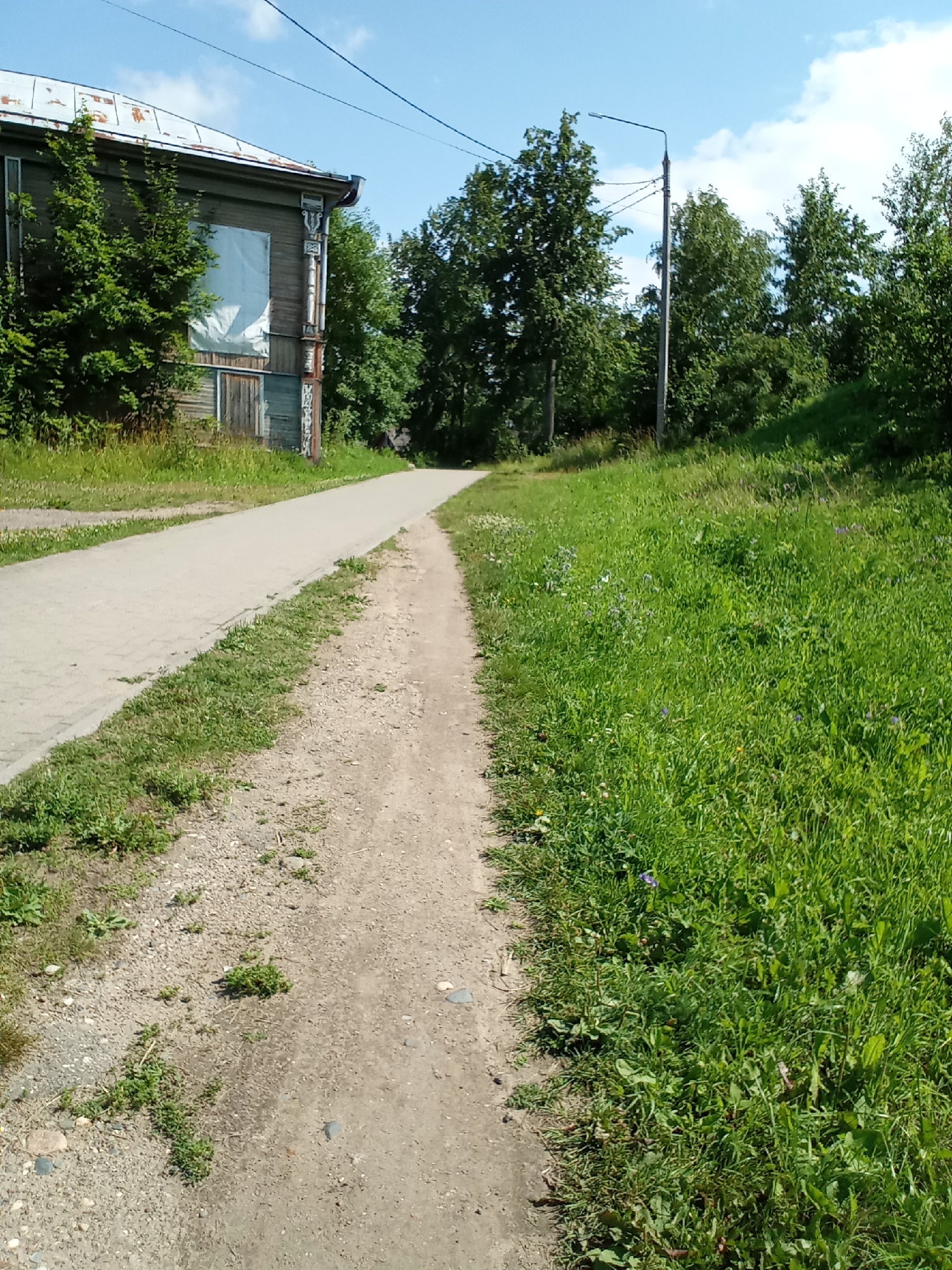 The image depicts a dirt path and a paved walkway alongside a grassy area with trees and a wooden building. The scene is outdoors and appears to be in a rural or suburban setting. In the background, a utility pole with a light fixture is visible, along with power lines. The wooden building, partially covered by what appears to be a tarp or sheeting over a window, has a weathered appearance. The paved walkway is adjacent to a grassy slope dotted with small wildflowers. The overall impression is one of a calm, sunlit day. Based on the provided context, this scene is located in the city of Belozersk, Russia.