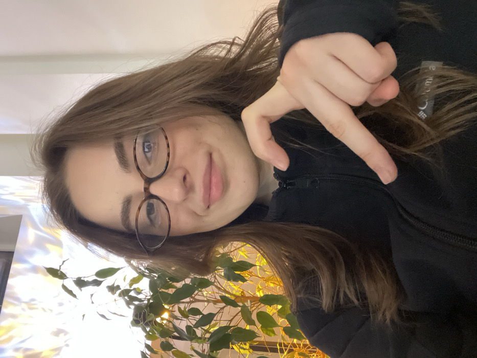 A young woman wearing glasses smiles slightly at the camera. She has long brown hair and wears a black Columbia jacket. Her hand is raised, making a "peace sign" or "victory" gesture. The background shows a wall partially obscured by a leafy plant. The light appears bright, casting a glow on the scene. The image is presented in a slightly rotated portrait orientation.