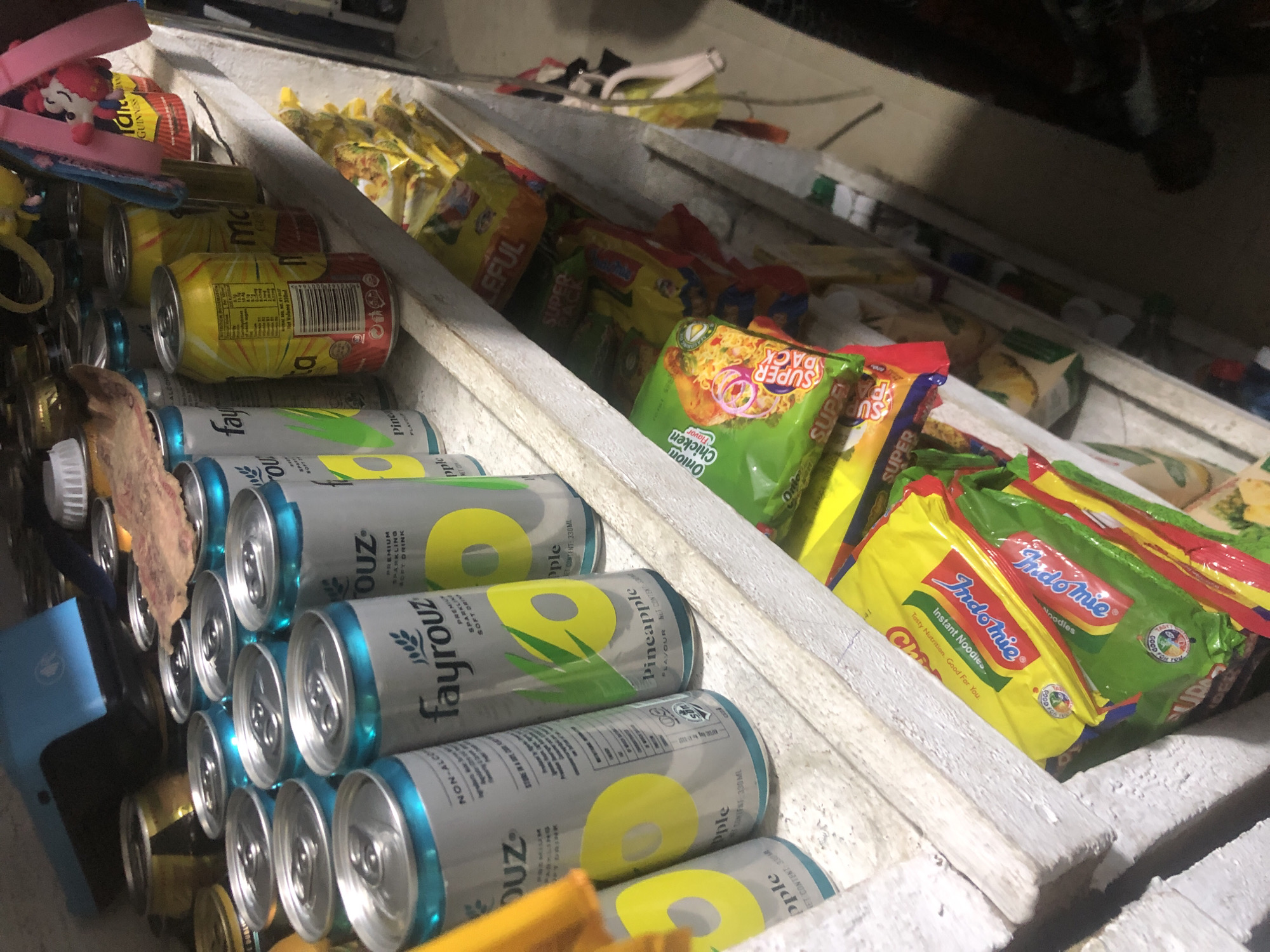This image shows a shelf in what appears to be a small convenience store or market stall in Ado, Nigeria. The shelf is stocked with various food and beverage items. Prominently displayed are several cans of Fayrouz sparkling soft drink, with prominent pineapple imagery. There are also packages of Indomie instant noodles, some in yellow packaging and others in green. Other items on the shelf include what appear to be more canned drinks, some with yellow and red packaging, and possibly other snack items. In the background, there are glimpses of other shelves stocked with various goods, and a blue plastic object that could be a cooler or storage container. The overall setting suggests a retail environment where these items are being sold.