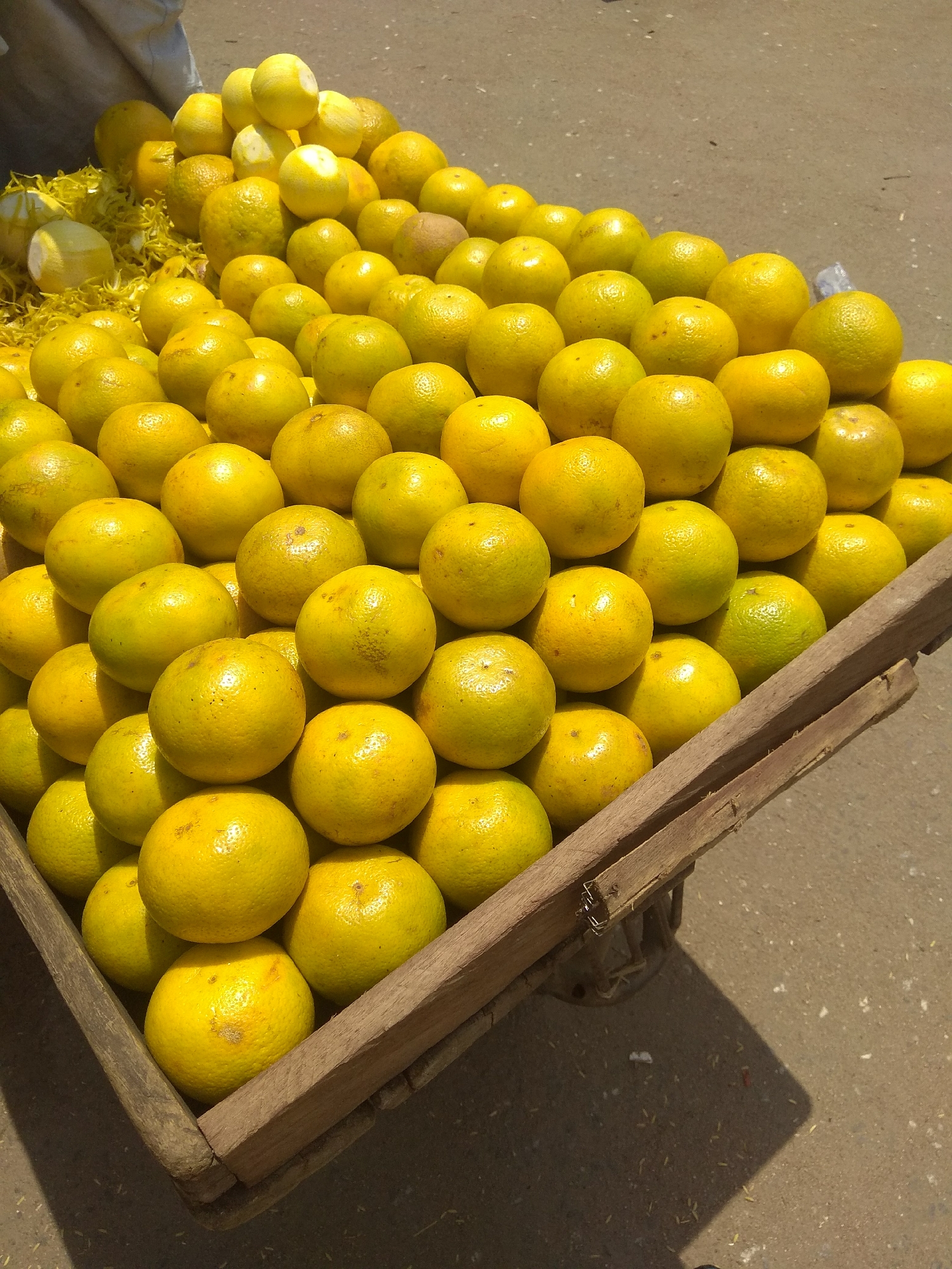 This image depicts a close-up, high-angle view of a wooden cart overflowing with ripe oranges, likely displayed for sale on a street in Jalingo, Nigeria. The cart, constructed from rough-hewn wooden planks, is filled to capacity with the citrus fruits, which are arranged in a dense, tiered fashion. The oranges are predominantly a vibrant yellow-orange, indicating their ripeness, and their surfaces show subtle variations in texture and color, with some exhibiting slight blemishes or lighter patches.
In the background, out of focus, a light-colored fabric, possibly a tarp or clothing, is visible, along with the grey asphalt of a street. The ground beneath the cart is also asphalt, scattered with small bits of debris, likely straw or chaff. The lighting suggests it is daytime, casting subtle shadows. No people are directly visible in the frame, but the presence of the cart filled with produce strongly implies a commercial transaction is occurring or about to occur in this urban environment. The scene is characteristic of a local market or roadside vendor in Jalingo.